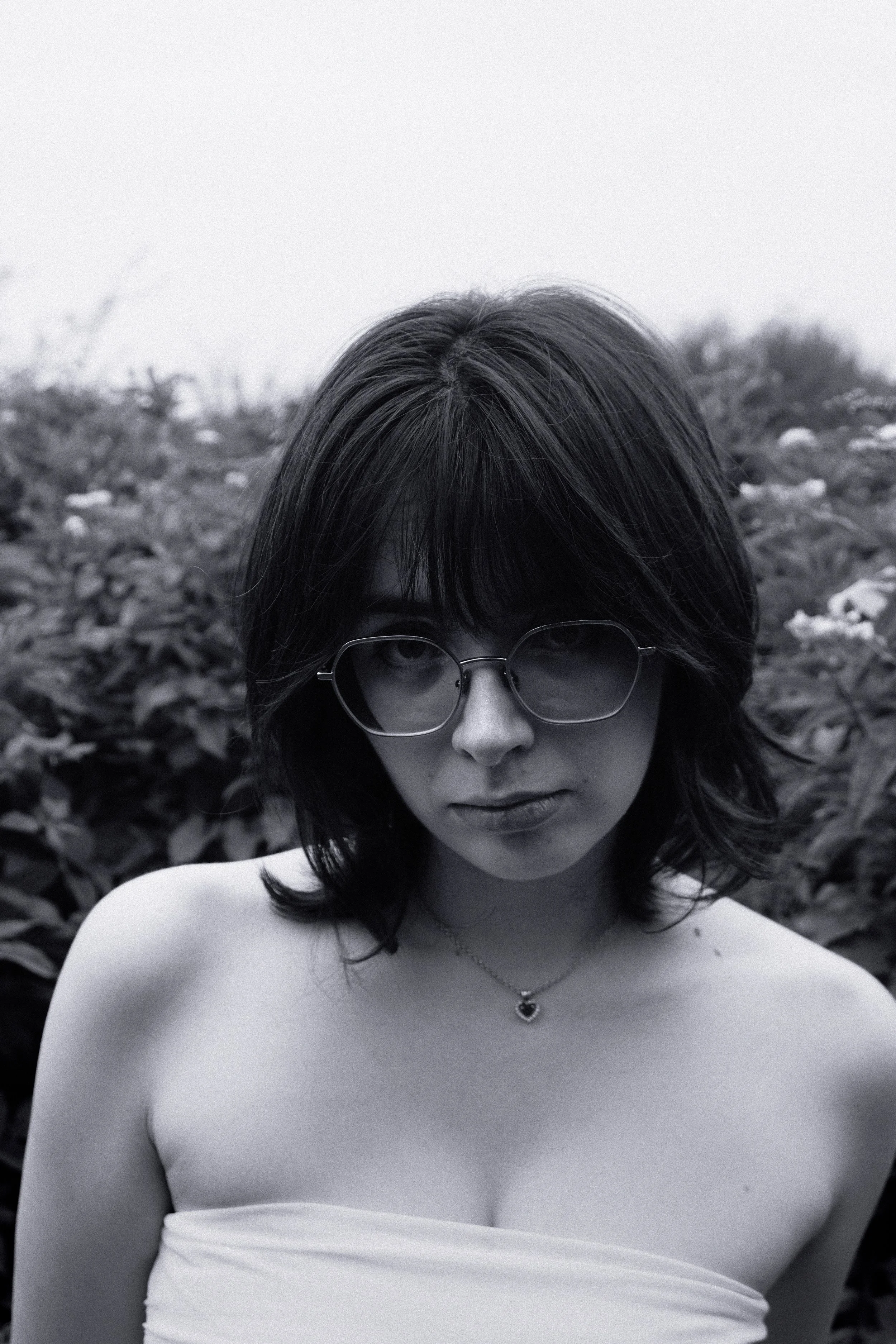 Jeune femme avec lunettes, cheveux longs et cheveux bruns, portant un collier en forme de cœur, en extérieur avec des feuilles en arrière-plan, en noir et blanc.