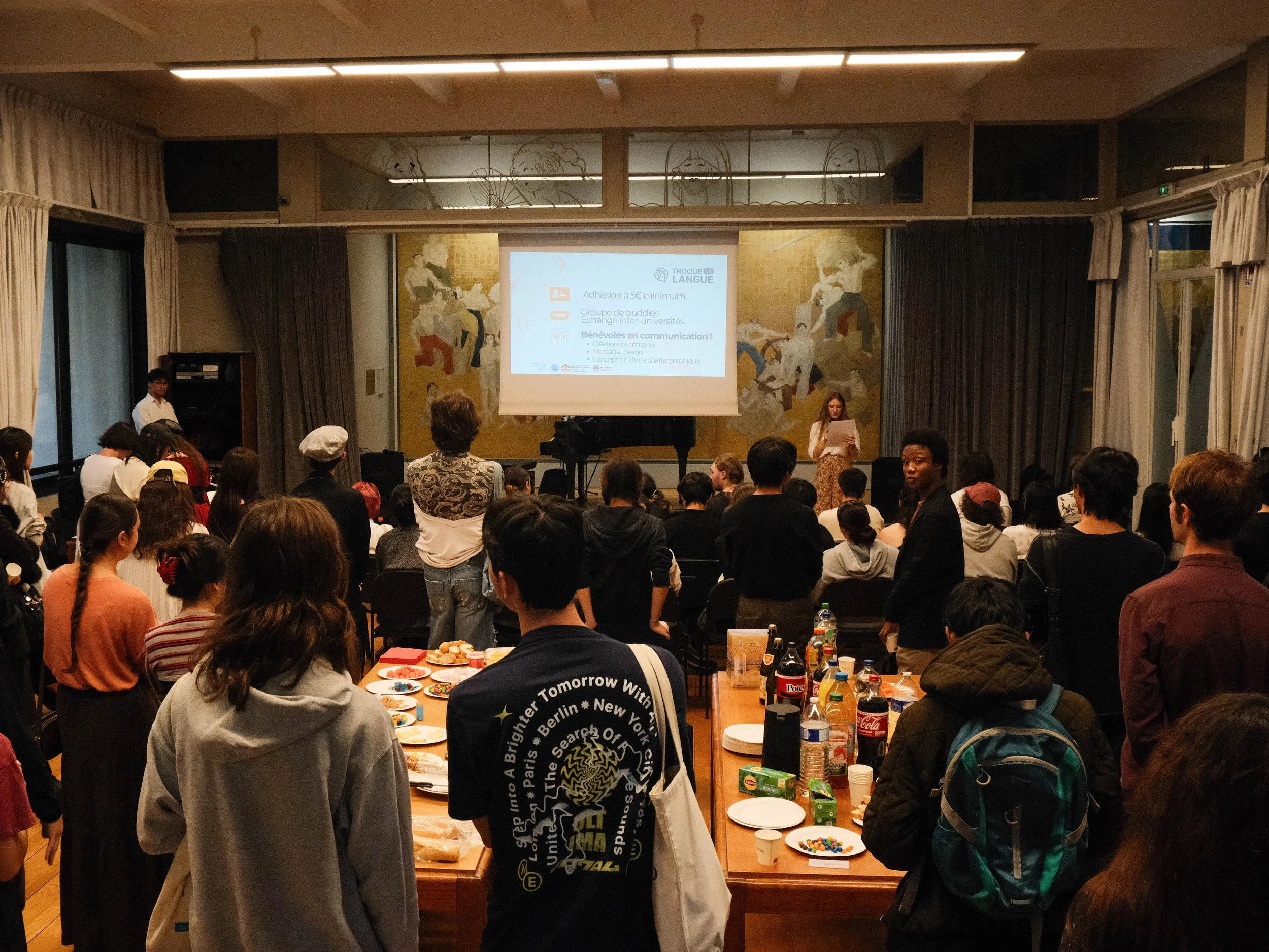 Une salle de conférence remplie de personnes assises ou debout, participant à un événement, avec une présentation projetée à l'avant de la pièce, et des tables avec des boissons et des snacks.