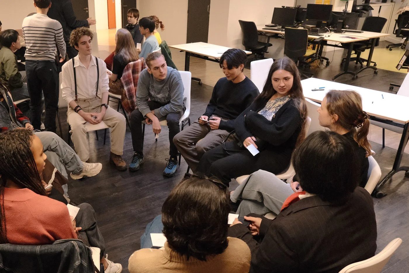 Un groupe de jeunes assis en cercle dans une salle de réunion ou de classe, engagés dans une discussion, avec plusieurs autres jeunes en arrière-plan.