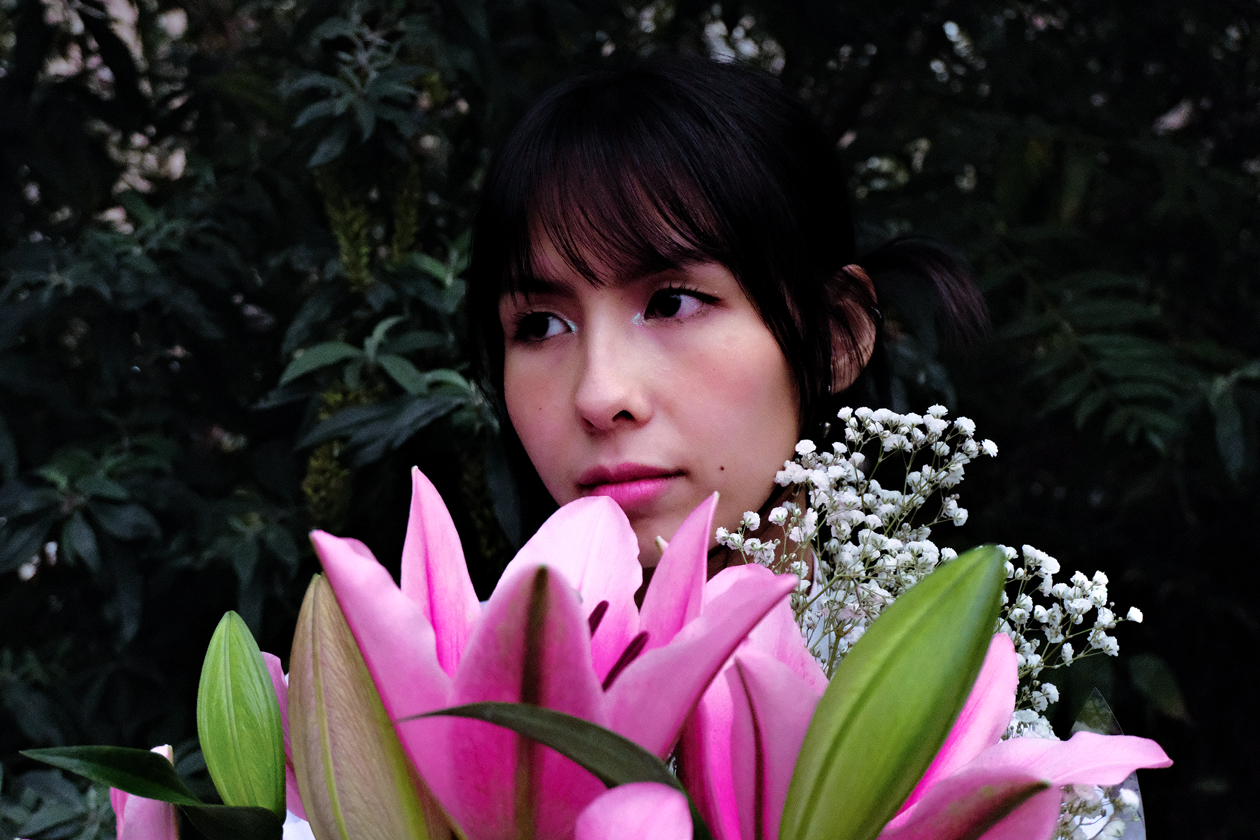 Une femme avec un visage délicat, coiffée de cheveux noirs avec une frange, entourée de fleurs roses, blanches et vertes dans un environnement naturel sombre.