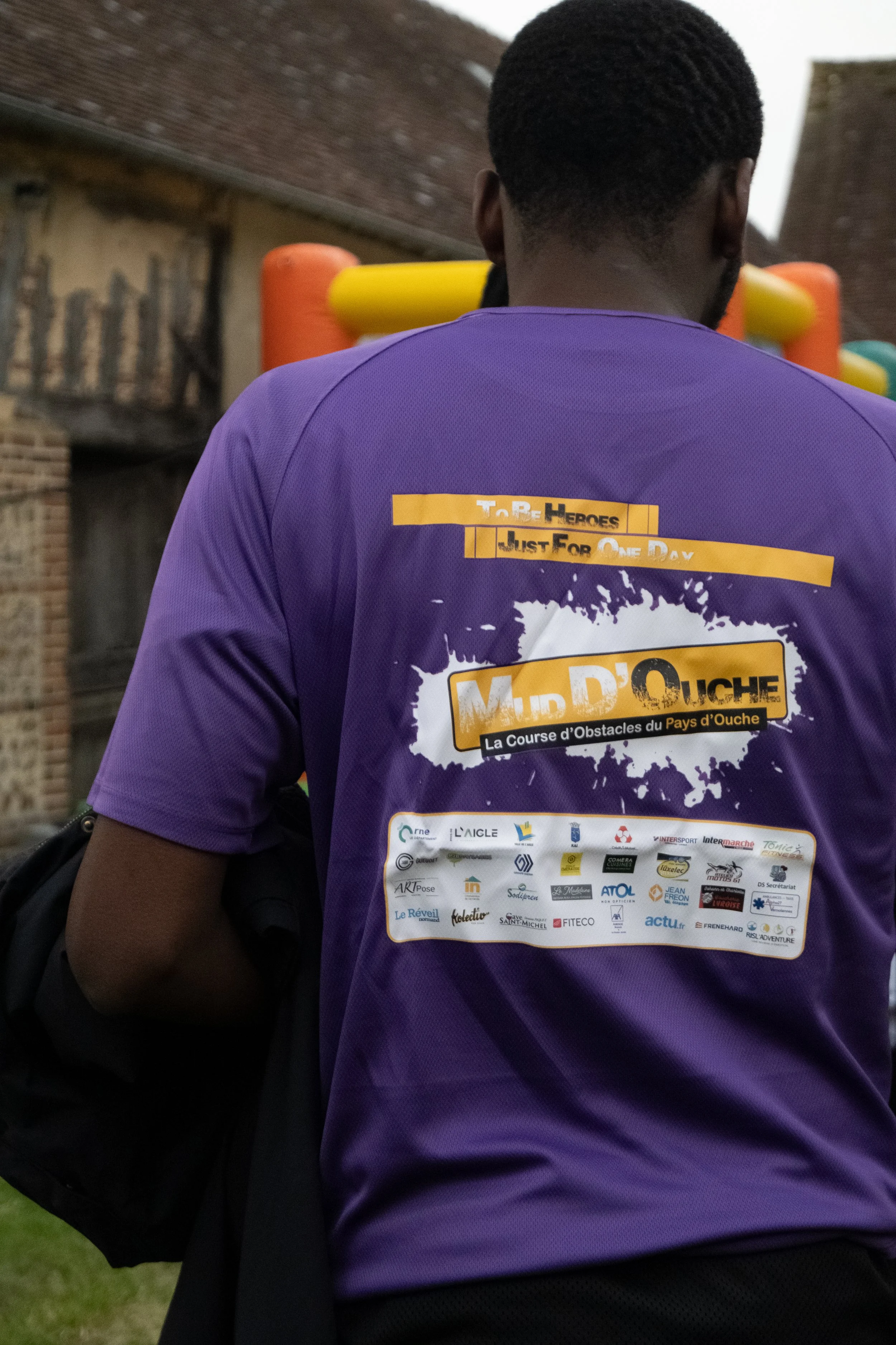 Personne portant un maillot violet avec des textes et logos, vue de dos, dans un espace extérieur, avec des bâtiments en briques en arrière-plan.