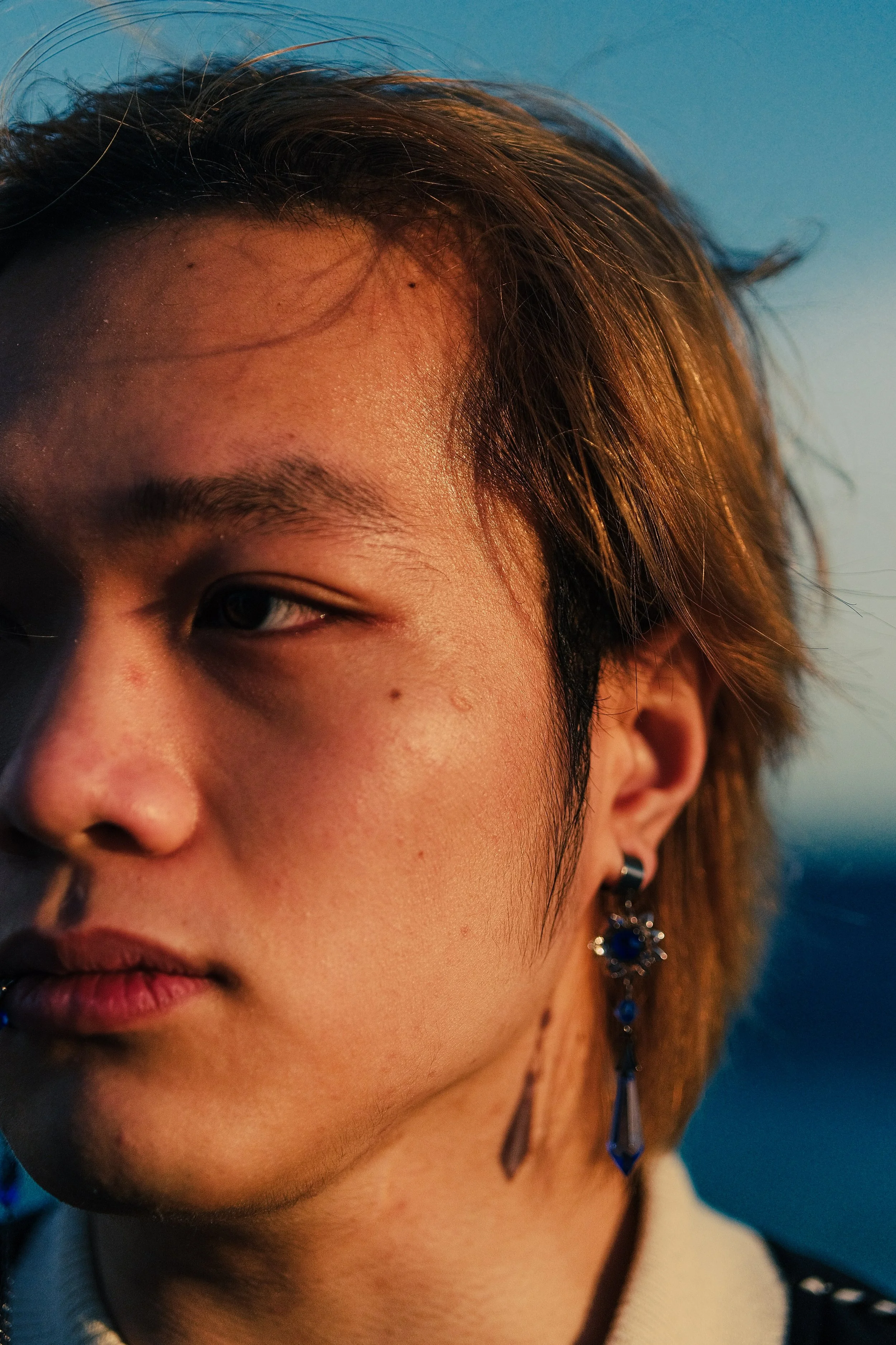 Gros plan d'une personne avec épaulement à gauche, portant une boucle d'oreille en forme de pierre précieuse bleue et des cheveux bruns. La personne a la peau claire et un fond bleu dégradé.