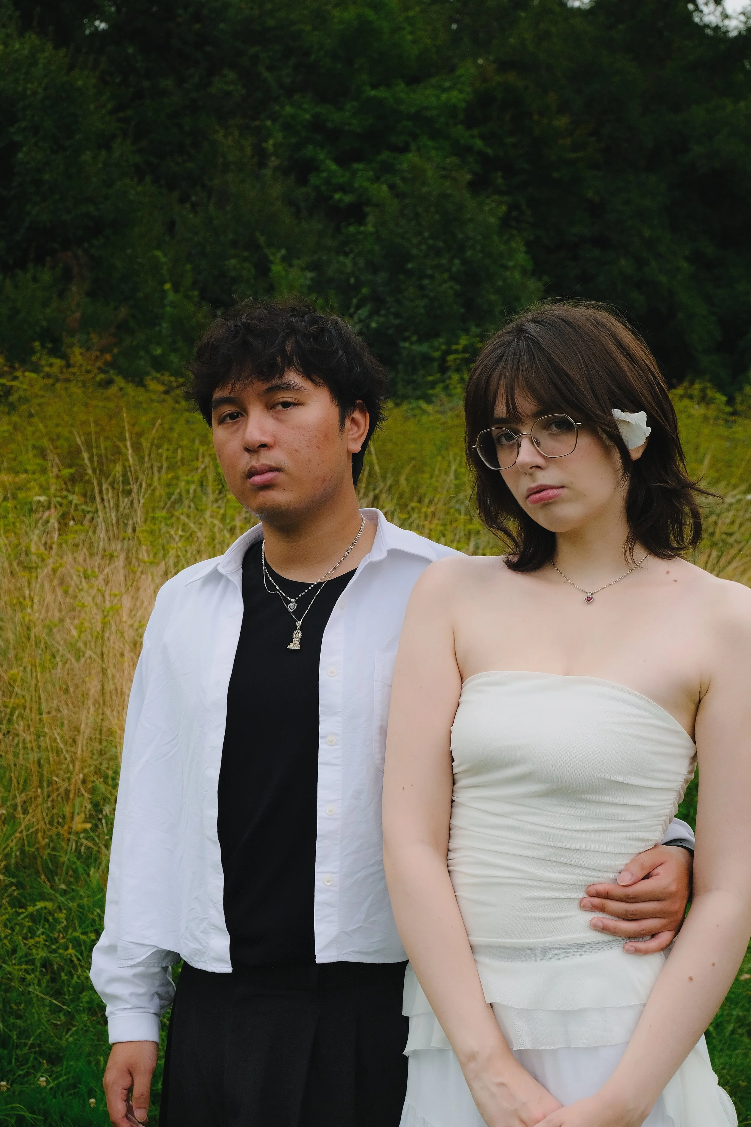 Deux jeunes adultes posant dans un champ avec un arrière-plan de forêt verte, l'homme porte une chemise blanche sur un t-shirt noir et des colliers, la femme porte une robe blanche strapless avec une fleur dans les cheveux et des lunettes.