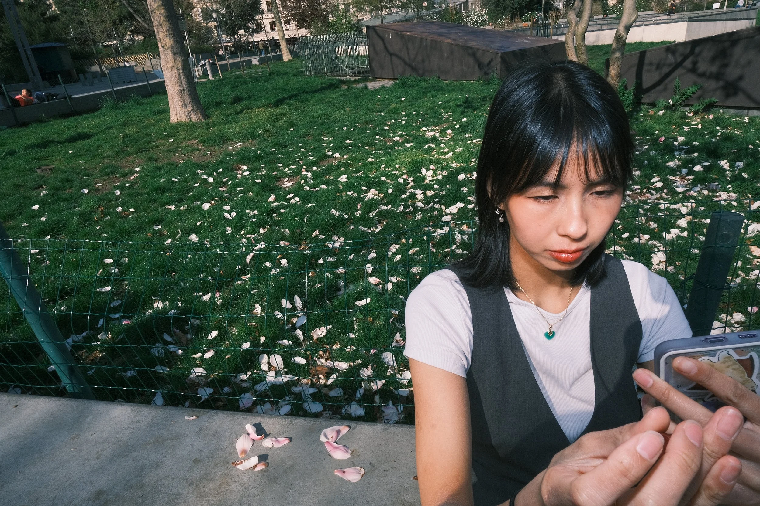 Une femme aux cheveux noirs courts, portant un t-shirt blanc et un gilet noir, regarde son téléphone, assise à côté d'une clôture en maillon de chaîne dans un parc avec un sol recouvert de pétales de fleurs. En arrière-plan, il y a des arbres et un b