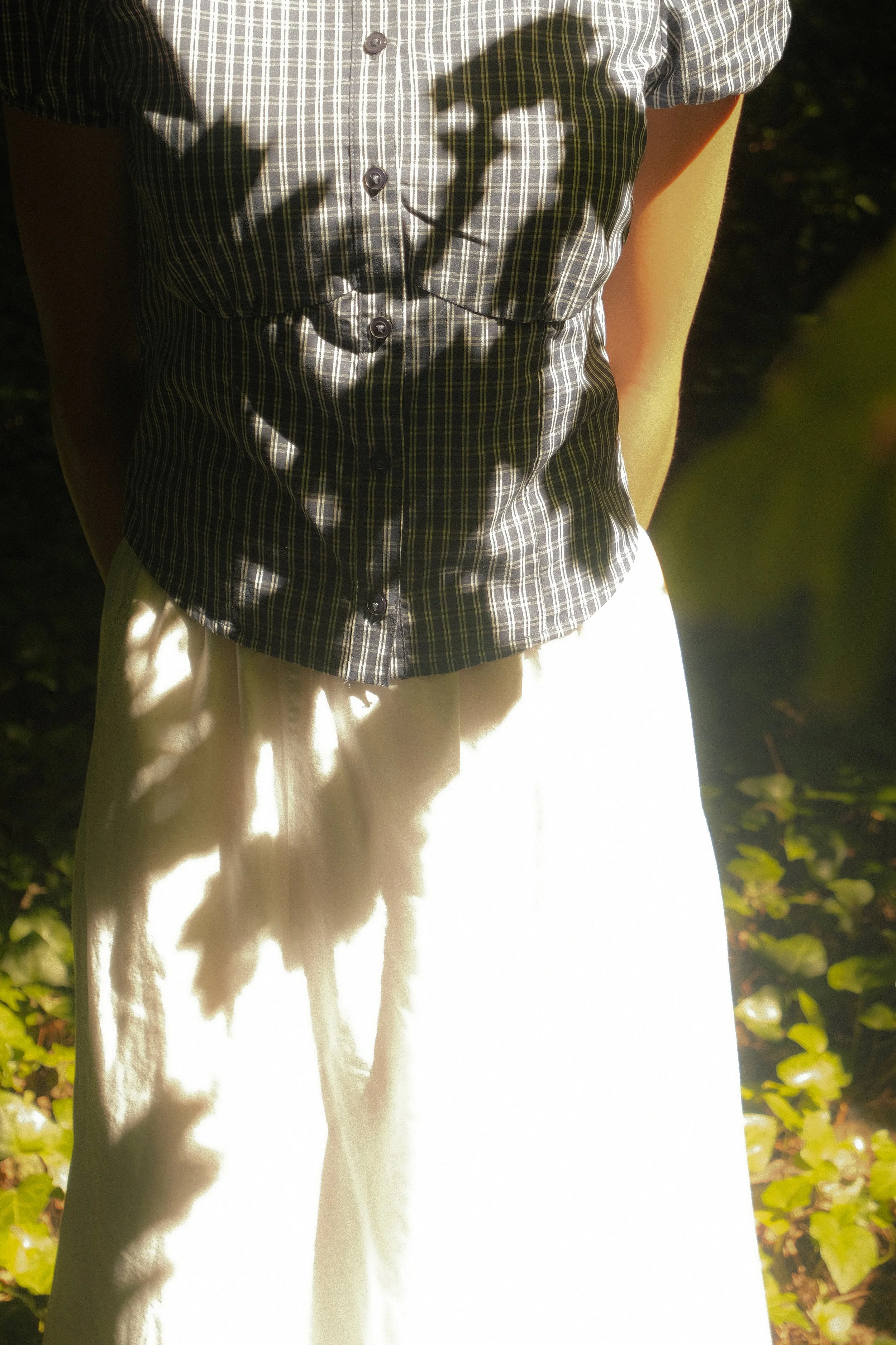 Personne portant une chemise à carreaux et une jupe blanche, debout dans la nature, avec des feuilles vertes autour.