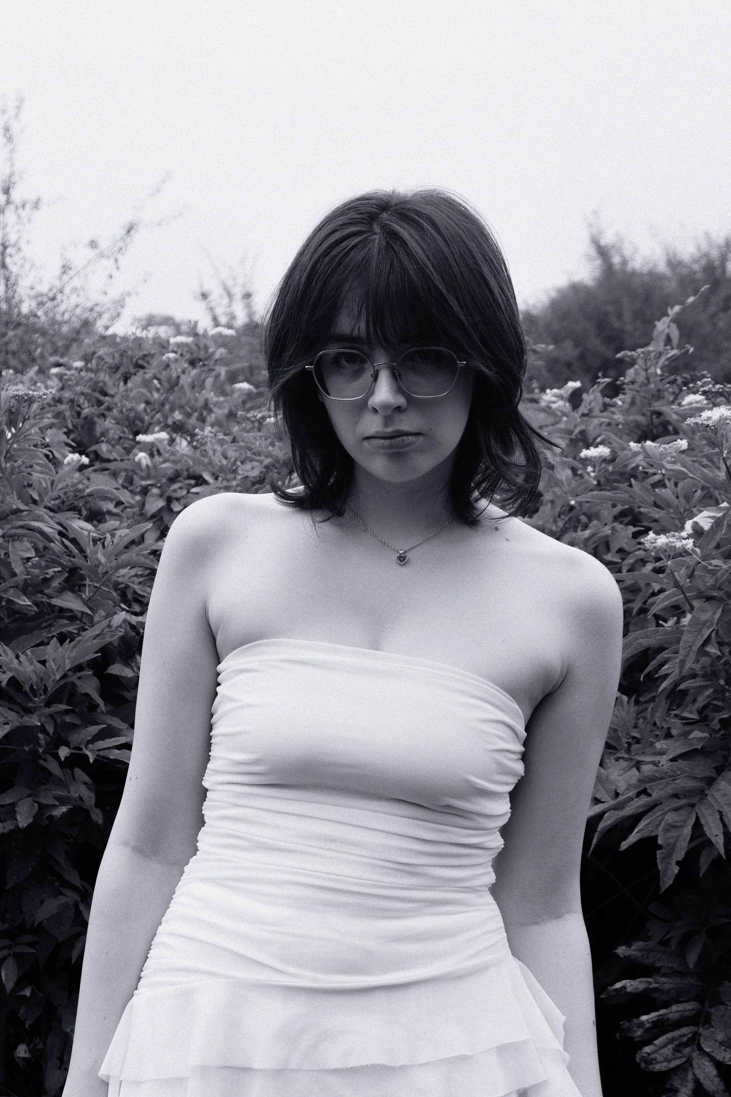 Une femme aux cheveux bruns, portant des lunettes rondes et un collier, en robe blanche dans un champ de plantes. Photo en noir et blanc.