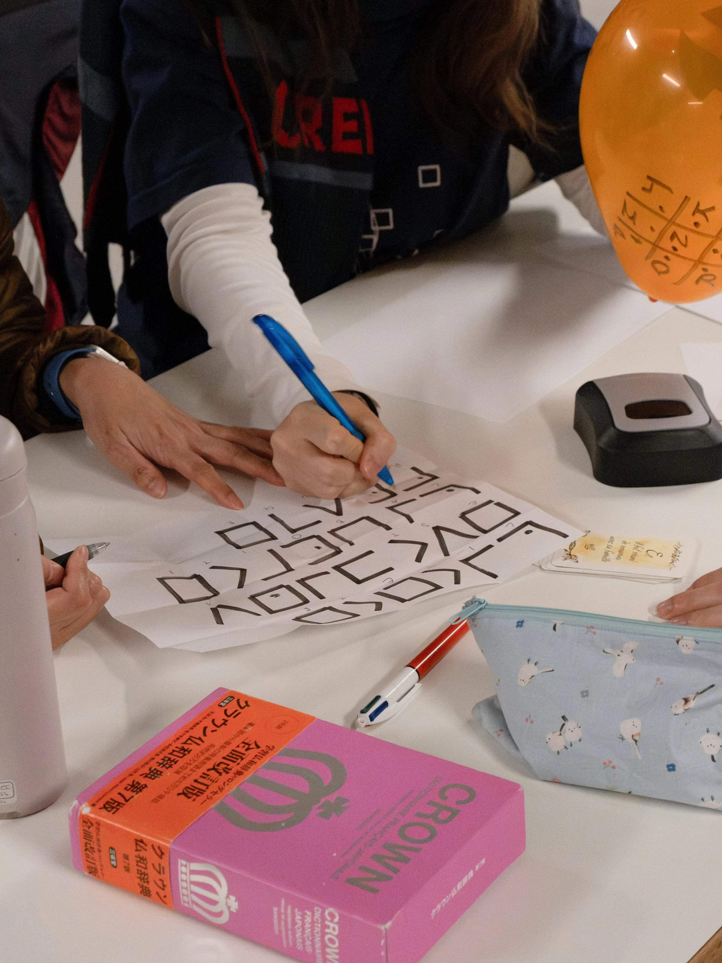 Personne dessinant des lettres de style graphique sur une feuille blanche, entourée d'objets de bureau, avec un ballon orange et une boîte rose sur la table.