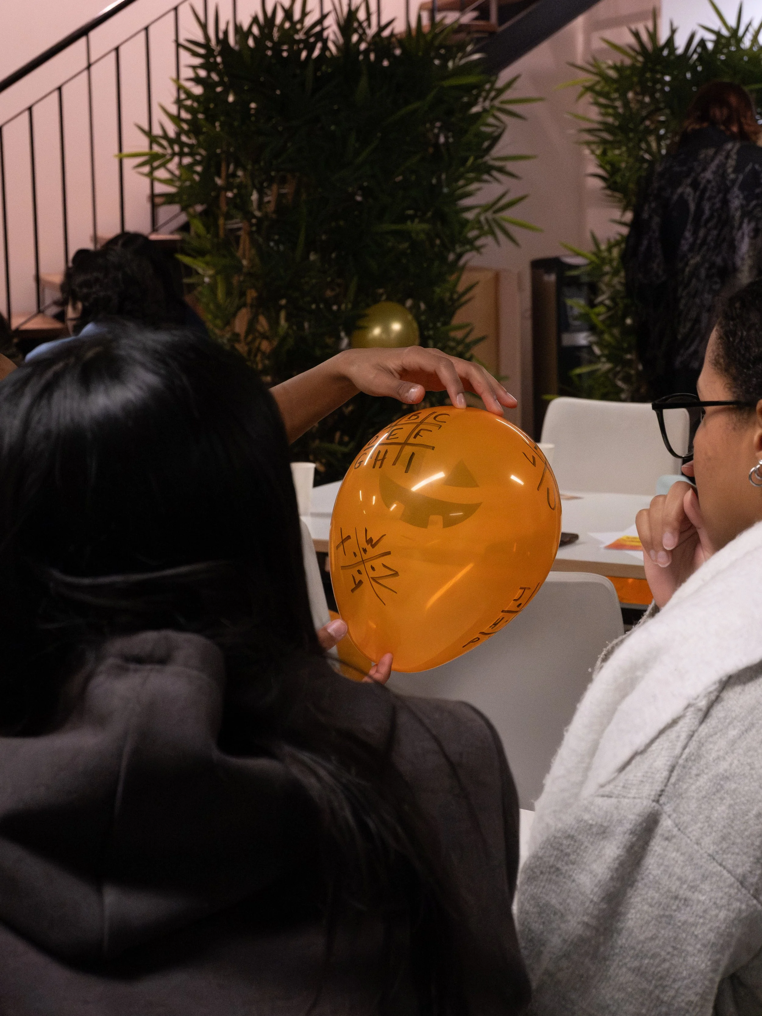 Deux personnes tiennent un ballon orange avec un visage de citrouille de Halloween et des écritures en chinois, lors d'une fête avec des plantes et des décorations de saison en arrière-plan.