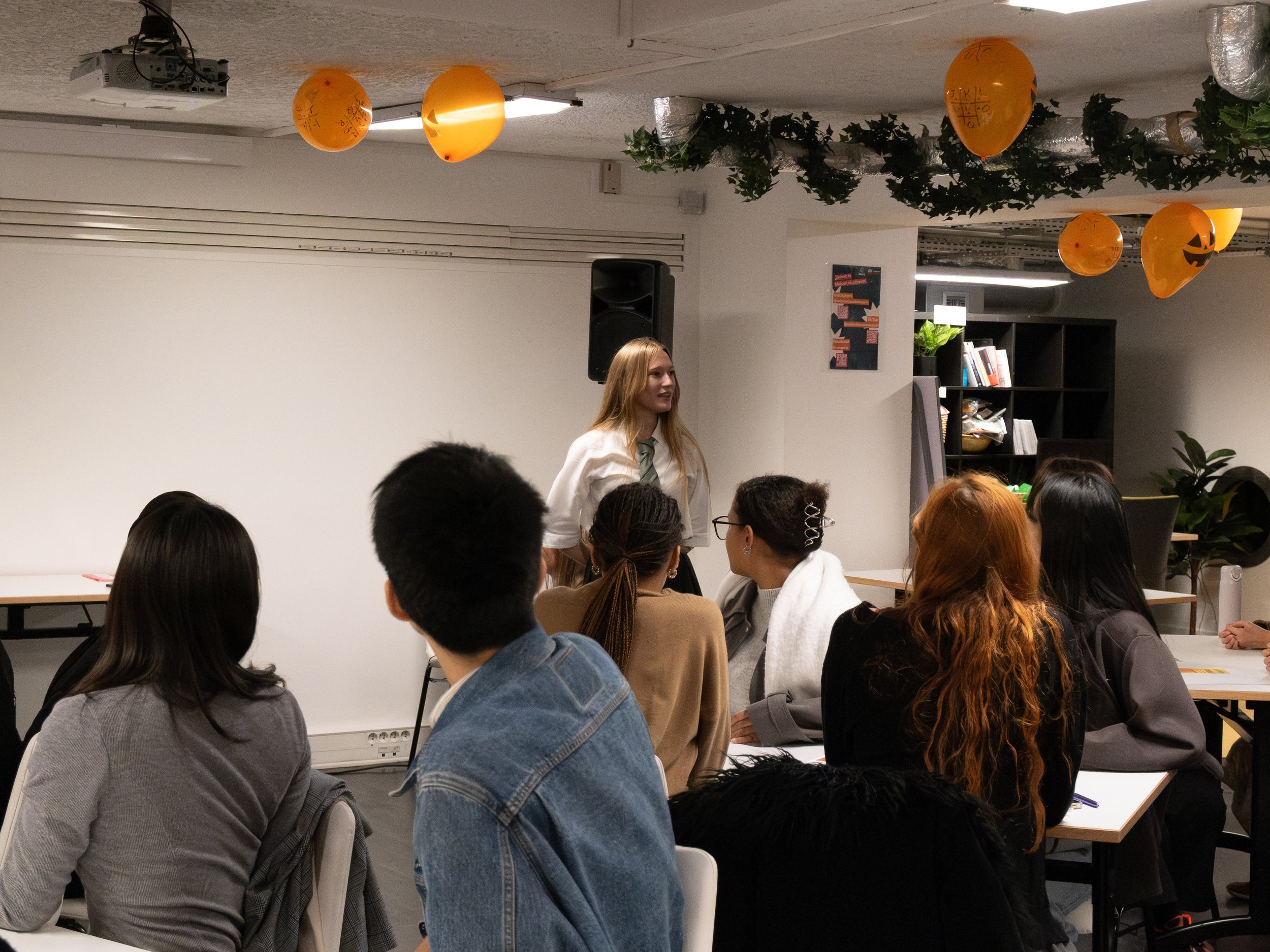 Une femme blonde en tenue blanche parlant devant un groupe de jeunes adultes dans une salle de classe ou de réunion décorée de ballons orange Halloween suspendus au plafond.