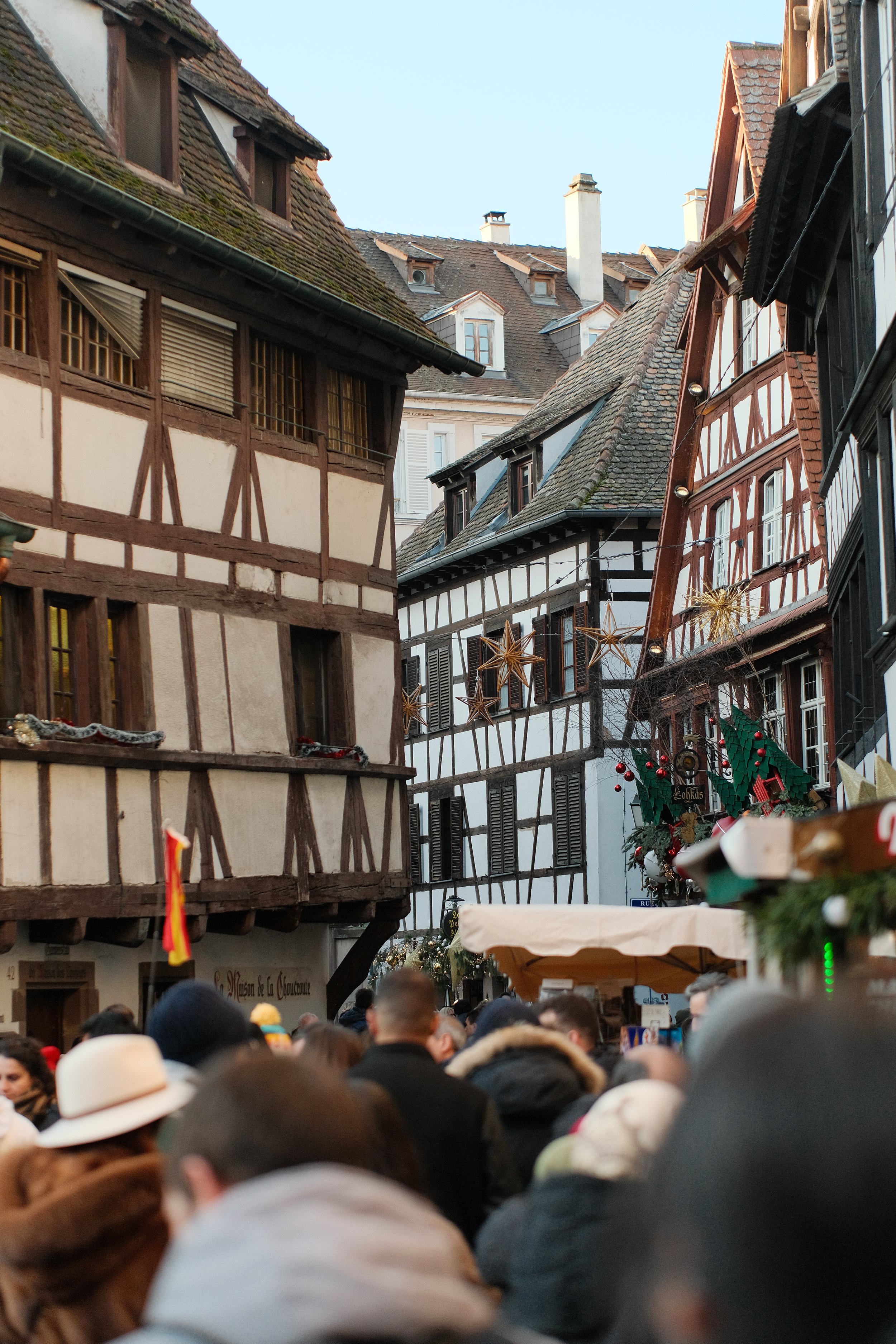 Une rue animée avec des bâtiments à colombages décorés pour Noël, des gens dans la foule, et des décorations de Noël suspendues dans les bâtiments.