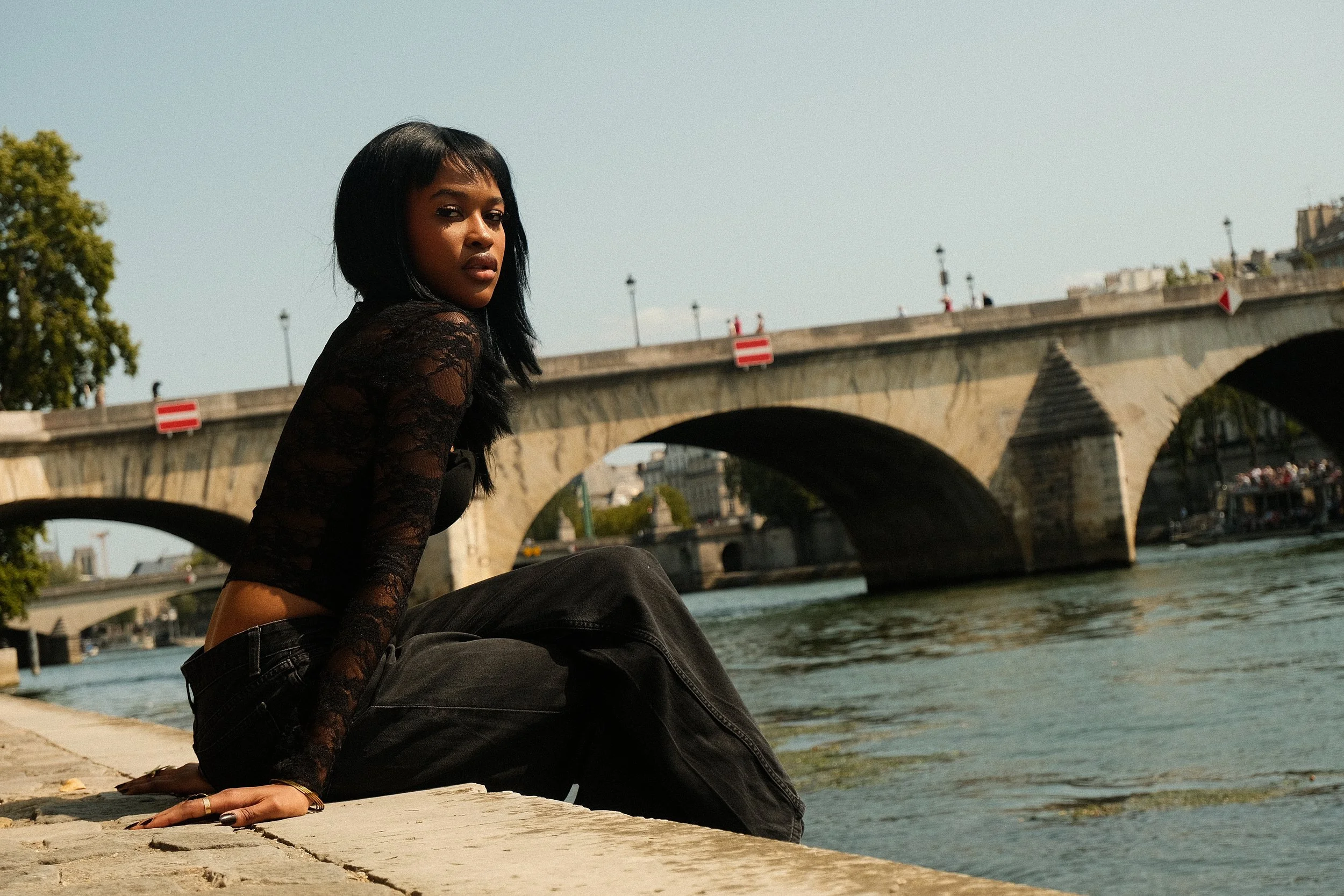 Une femme noire avec des cheveux noirs, portant un haut en dentelle noire et un pantalon noir, assise sur le bord de la Seine, avec un pont parisien en arrière-plan un jour ensoleillé.