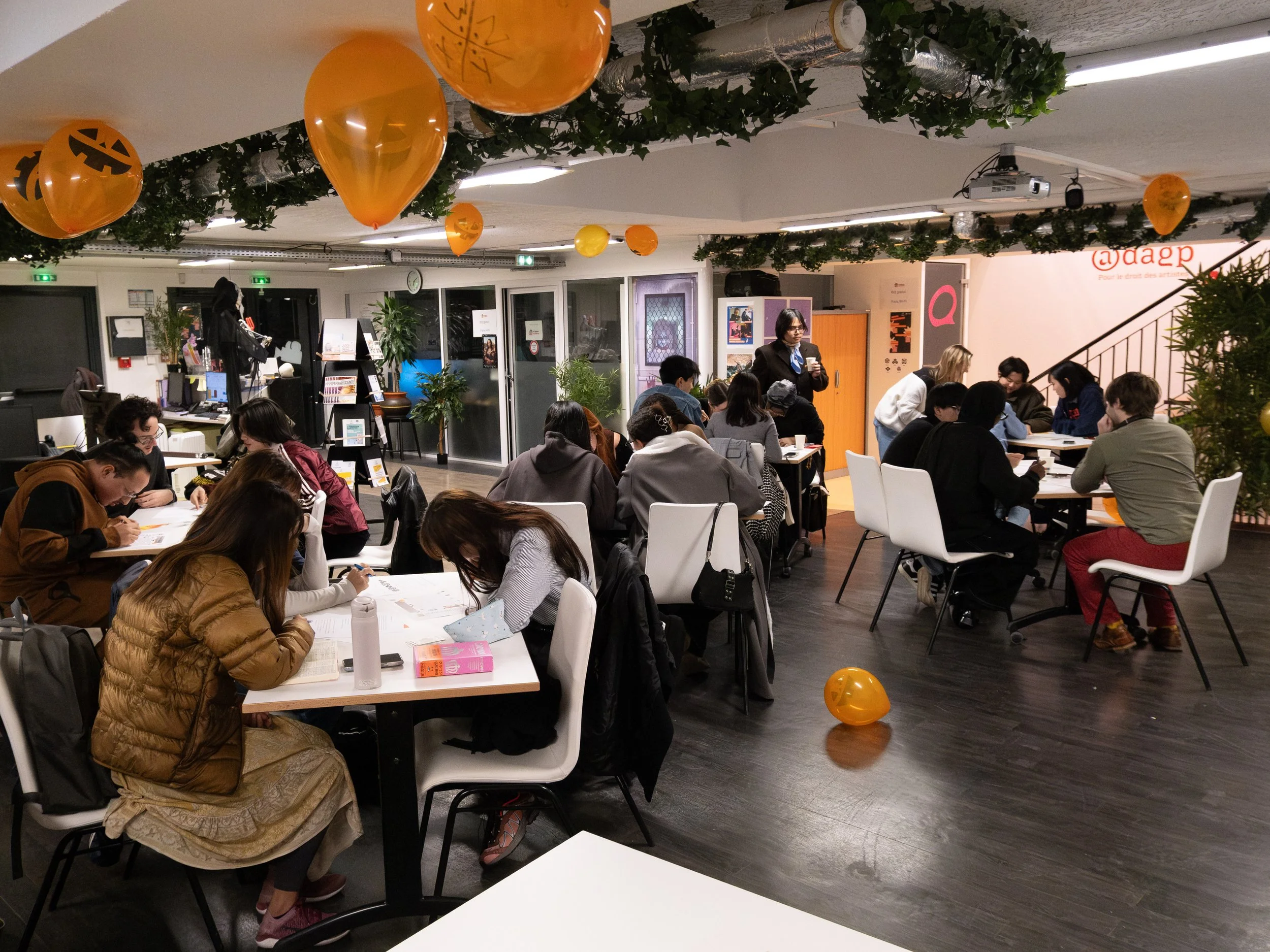 Une salle occupée par plusieurs groupes de personnes qui étudient ou travaillent, décorée avec des ballons orange Halloween suspendus au plafond, et des plantes vertes, dans un espace moderne avec des murs blancs et un coin cabine à l'arrière.