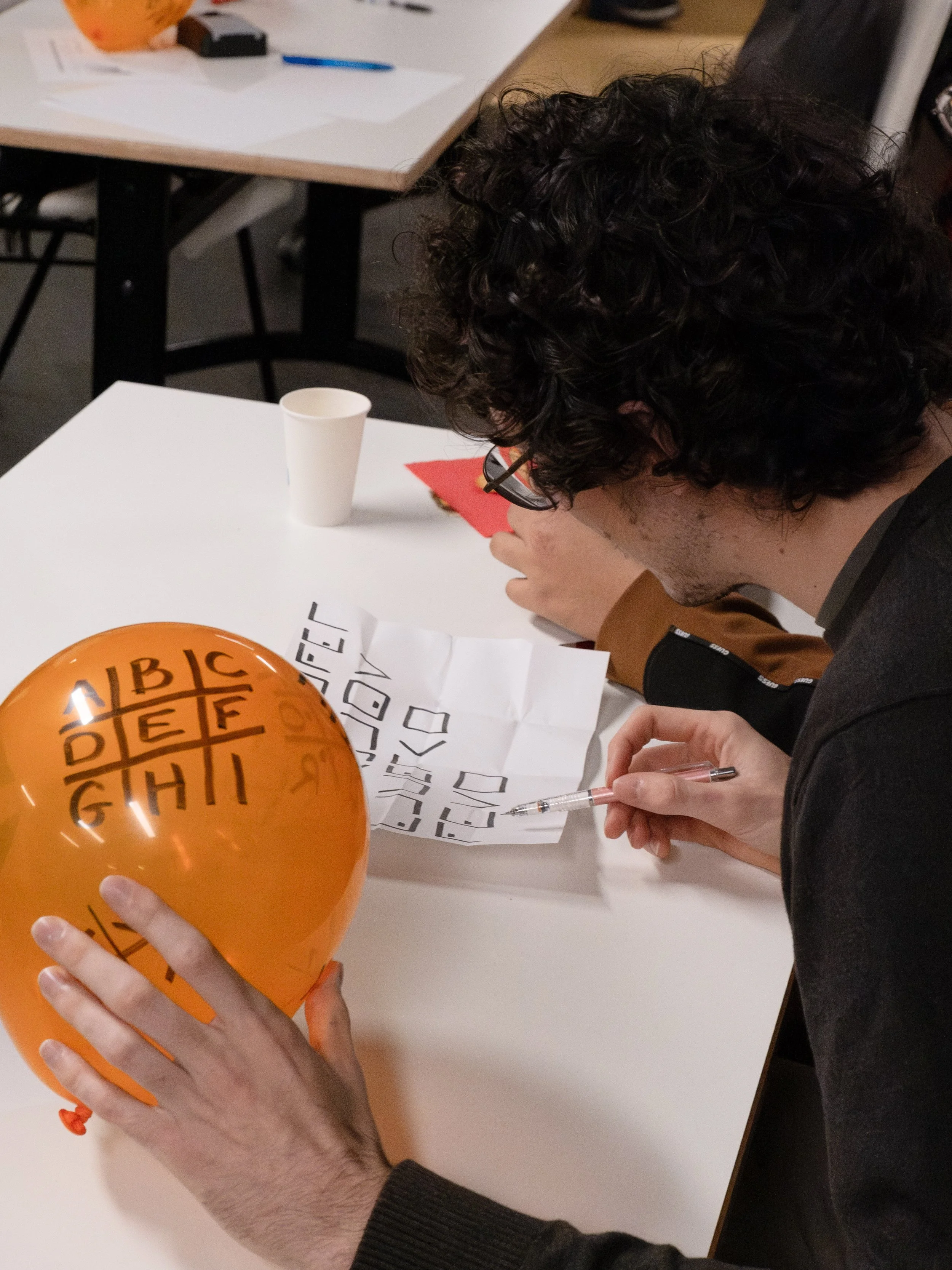 Une personne qui écrit à la main sur une feuille de papier avec un stylo, tenant un ballon orange avec des lettres noires, dans un environnement intérieur.