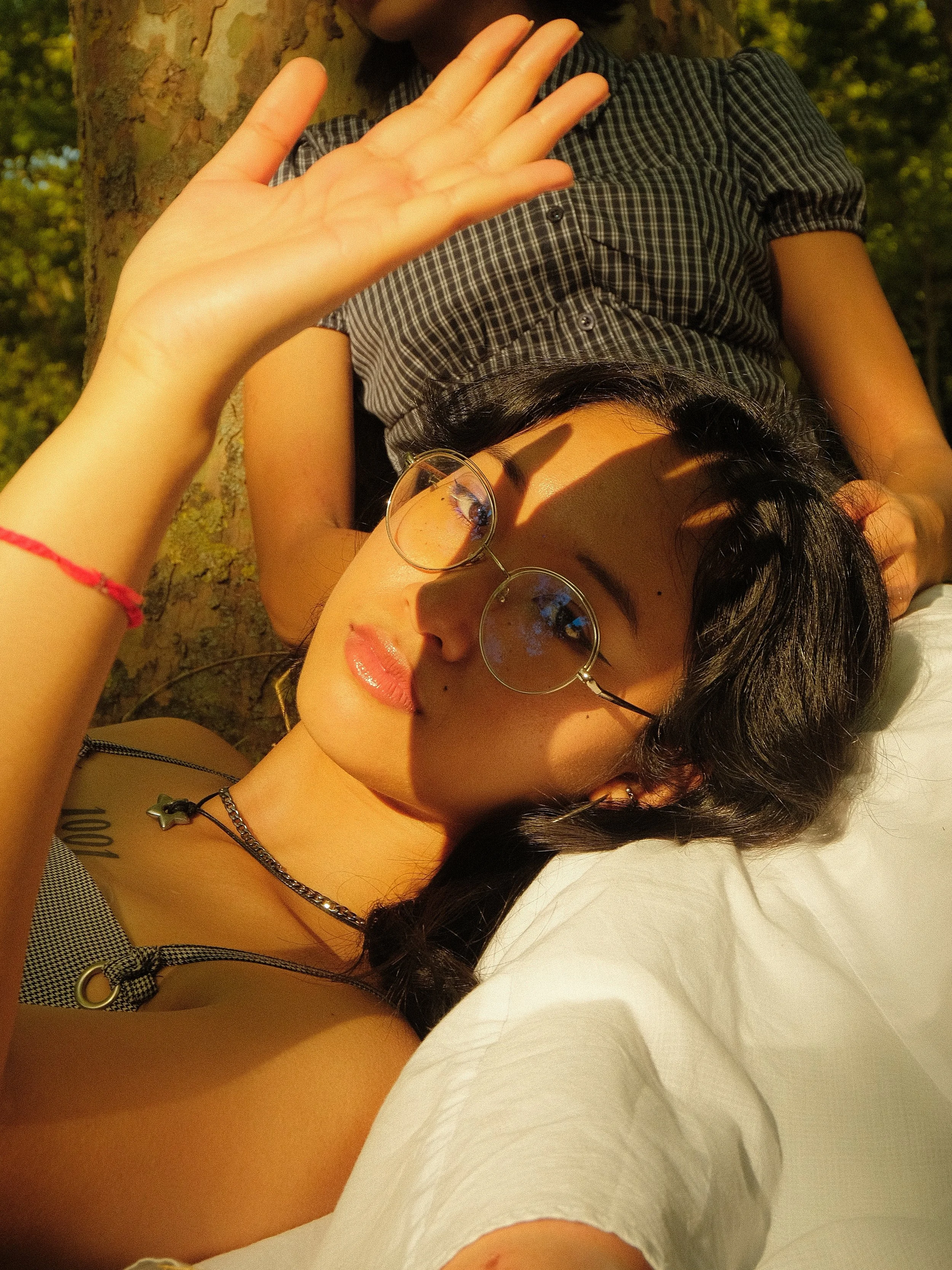 Une femme portant des lunettes rondes, allongée sur un lit ou un matelas à l'extérieur, avec une personne derrière elle dont seule une partie du corps est visible. La femme fait un signe de la main vers la caméra, en plein soleil, avec un arbre en ar