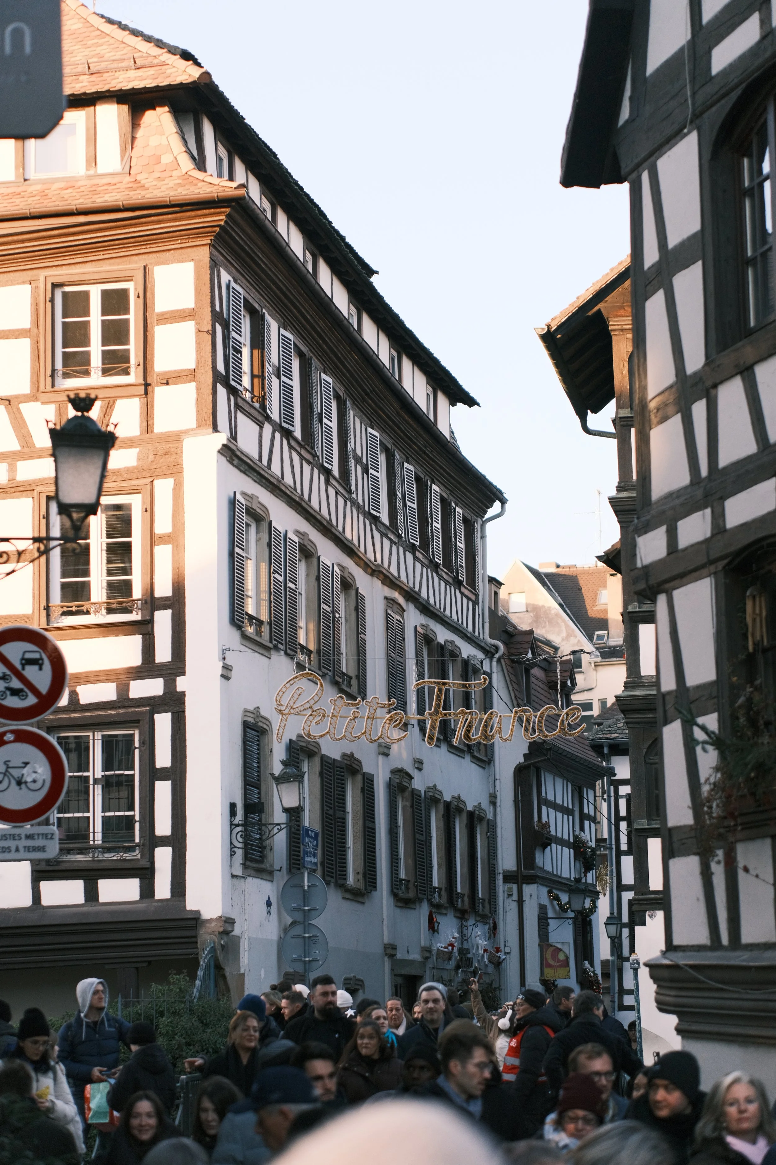 Une rue pavée de style alsacien avec des bâtiments à colombages et des fenêtres à volets, remplie de personnes, avec un signe lumineux 'Petite France' suspendu au centre.