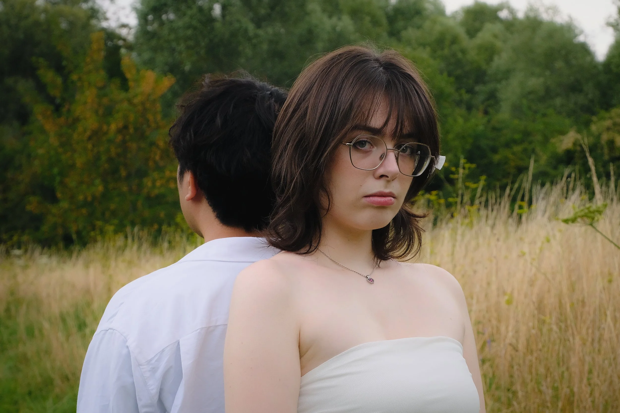 Une jeune femme avec des lunettes et des cheveux bruns, portant un haut blanc, se tient dans un champ d'herbes hautes avec un homme dont le dos est visible, dans un environnement naturel avec des arbres en arrière-plan.