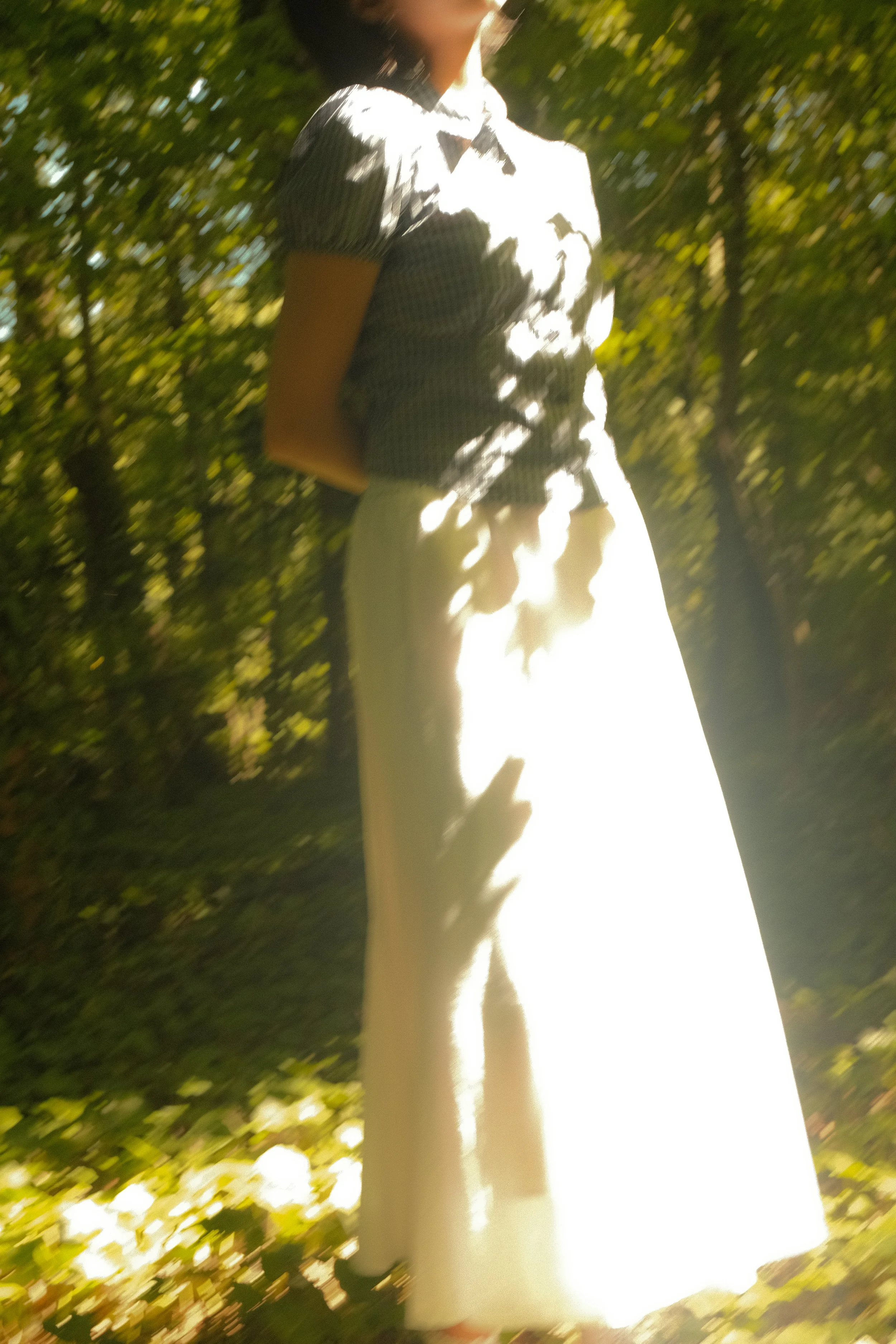 Une personne en robe longue blanche, debout dans une forêt, avec la lumière du soleil perçant à travers les arbres.
