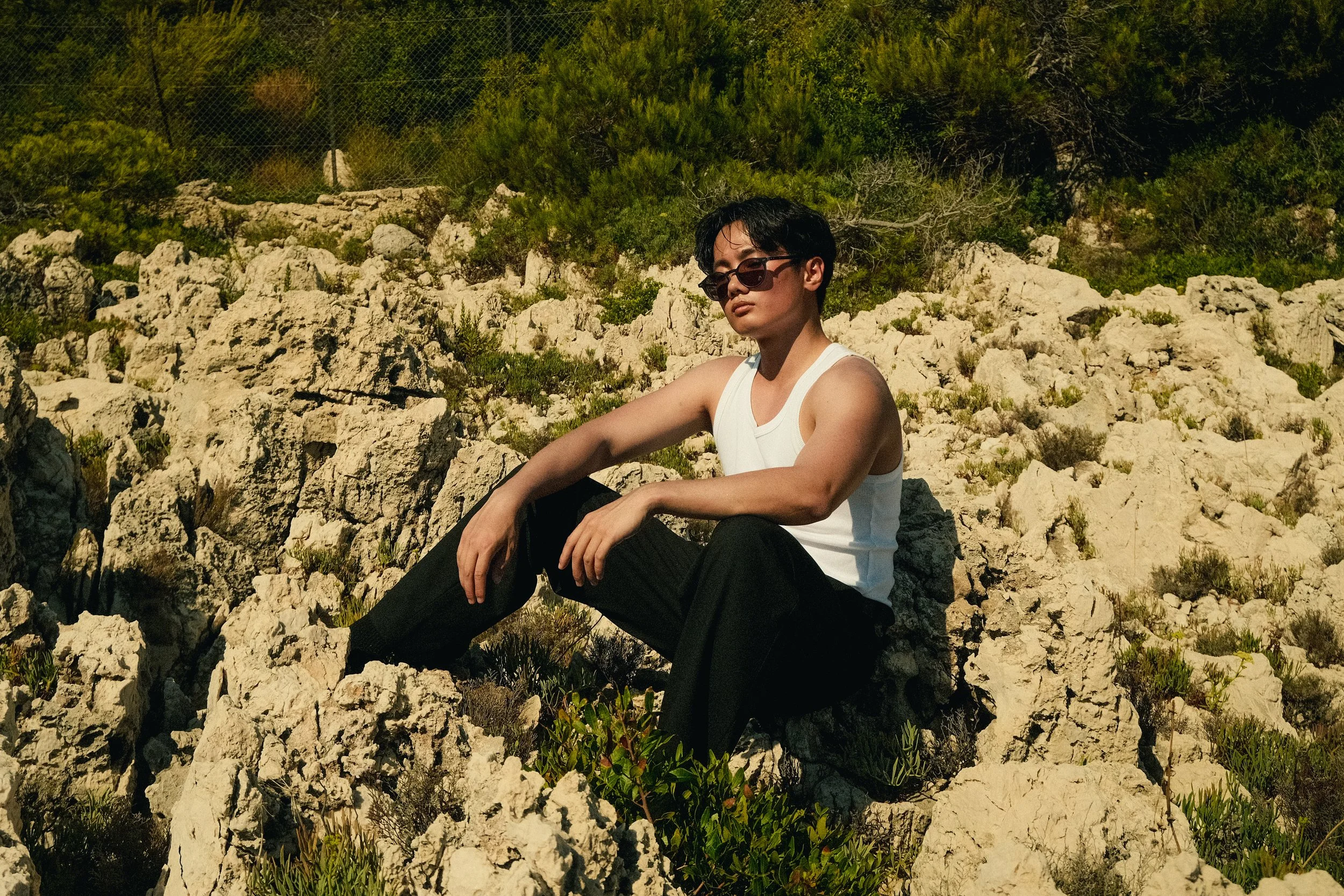 Une personne assise sur des rochers dans un environnement naturel en plein soleil, portant un débardeur blanc, des lunettes de soleil et des pantalons noirs.