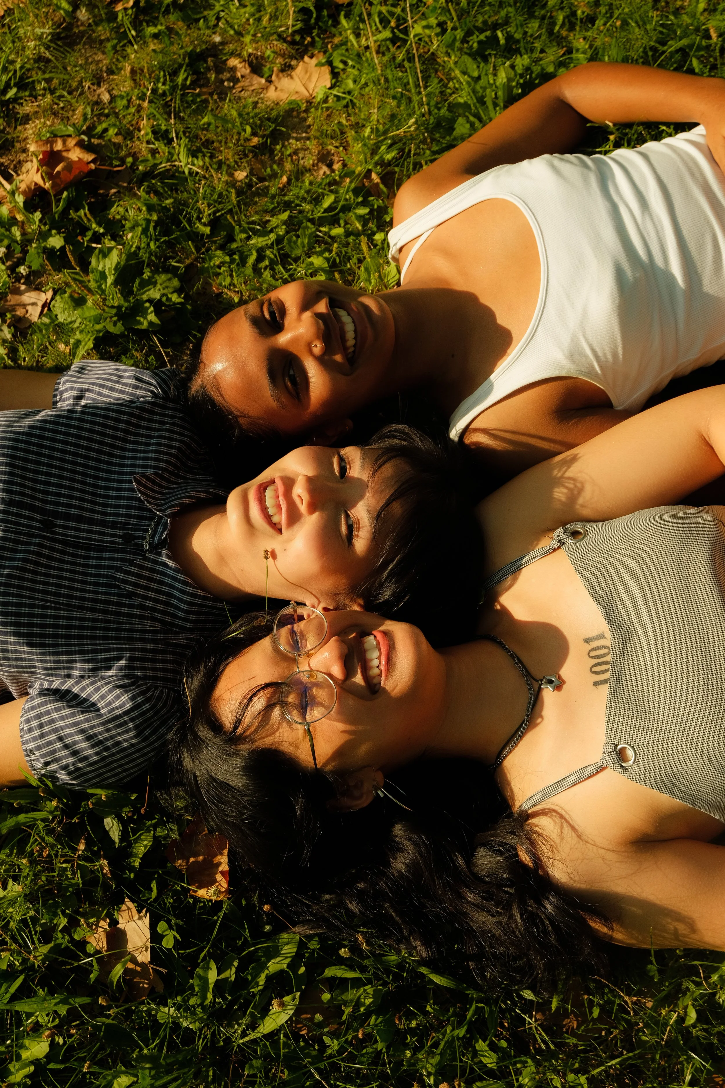 Trois femmes allongées dans l'herbe, souriantes, sous la lumière du soleil, en train de prendre une photo ou de se détendre ensemble.