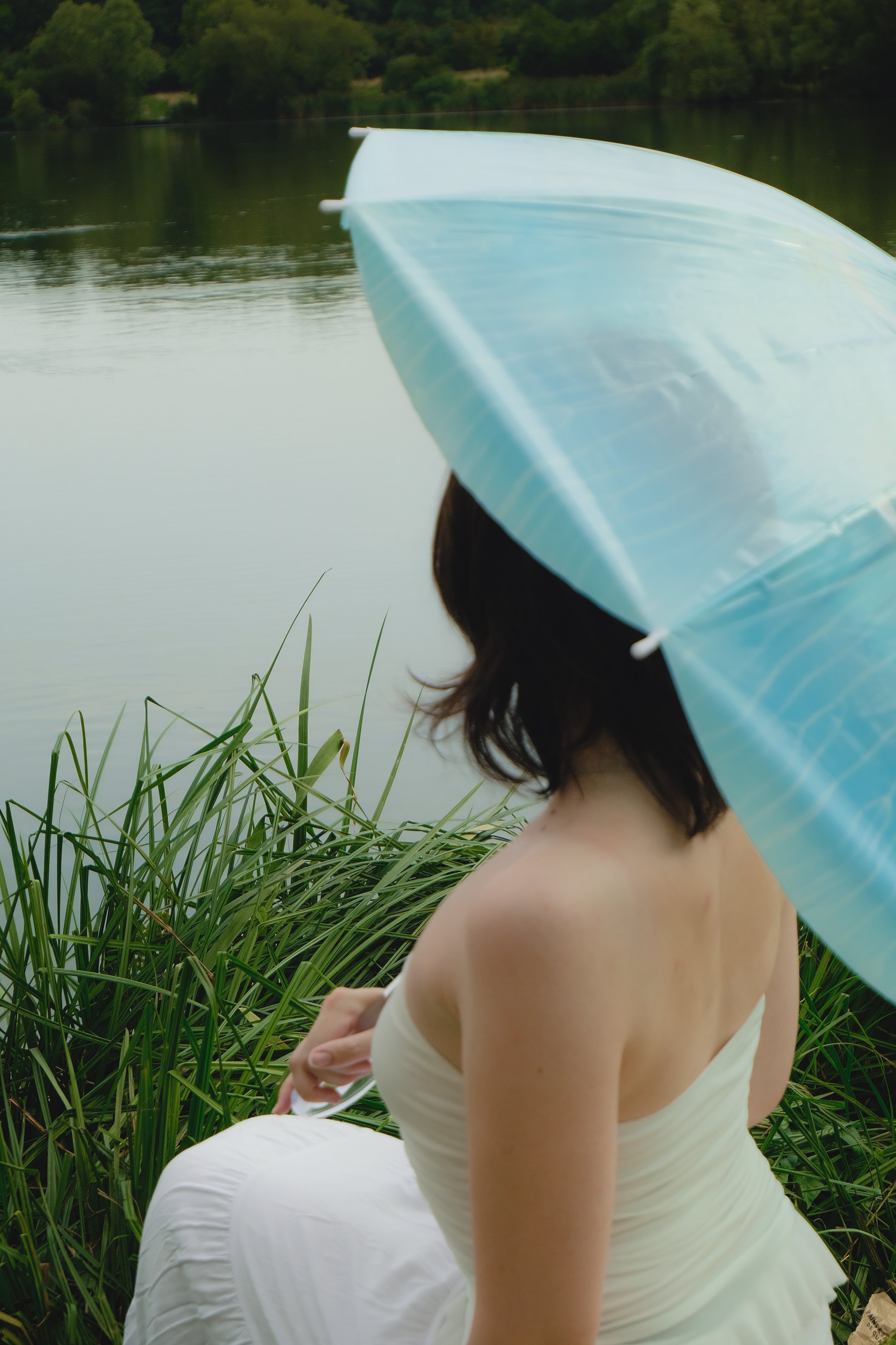 Une femme avec une robe blanche tient un parapluie bleu clair près d'un lac entouré de végétation.