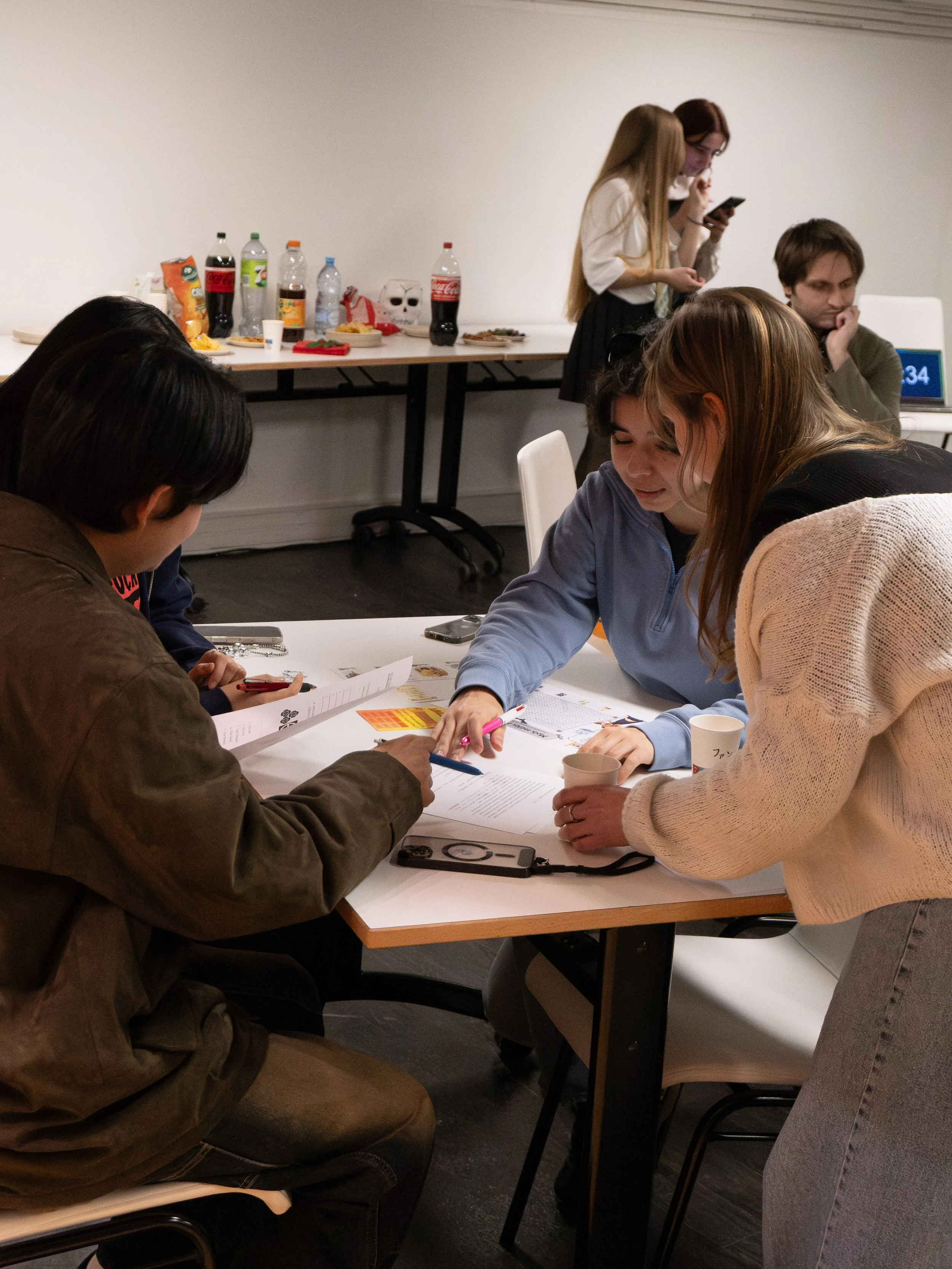 Groupe de jeunes adultes discutant autour d'une table, avec d'autres personnes en arrière-plan, une table avec des boissons et snacks.