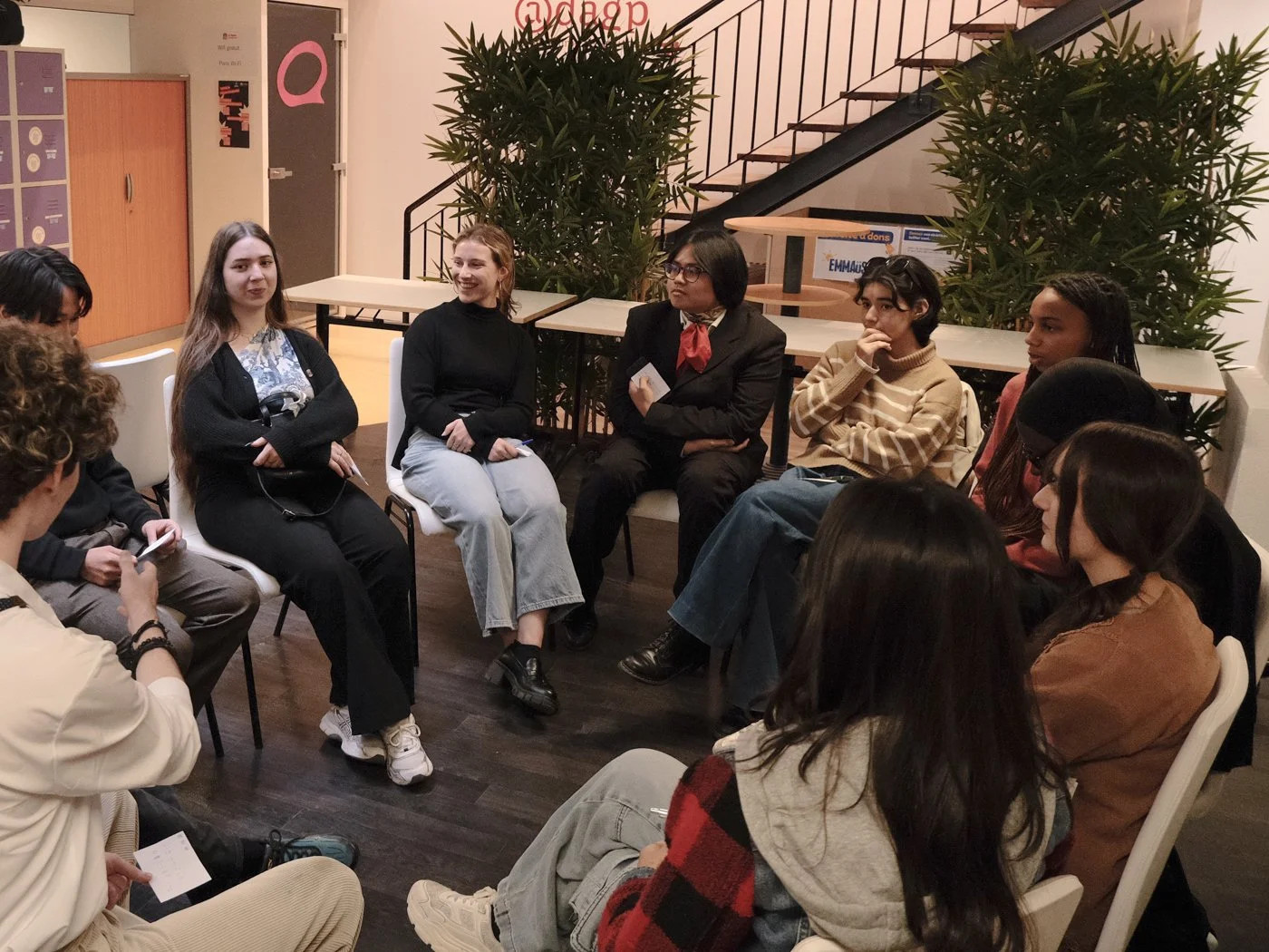 Groupe de jeunes femmes et hommes assis en cercle dans une salle, participant à une discussion ou un atelier, avec zones d'arbustes en arrière-plan.