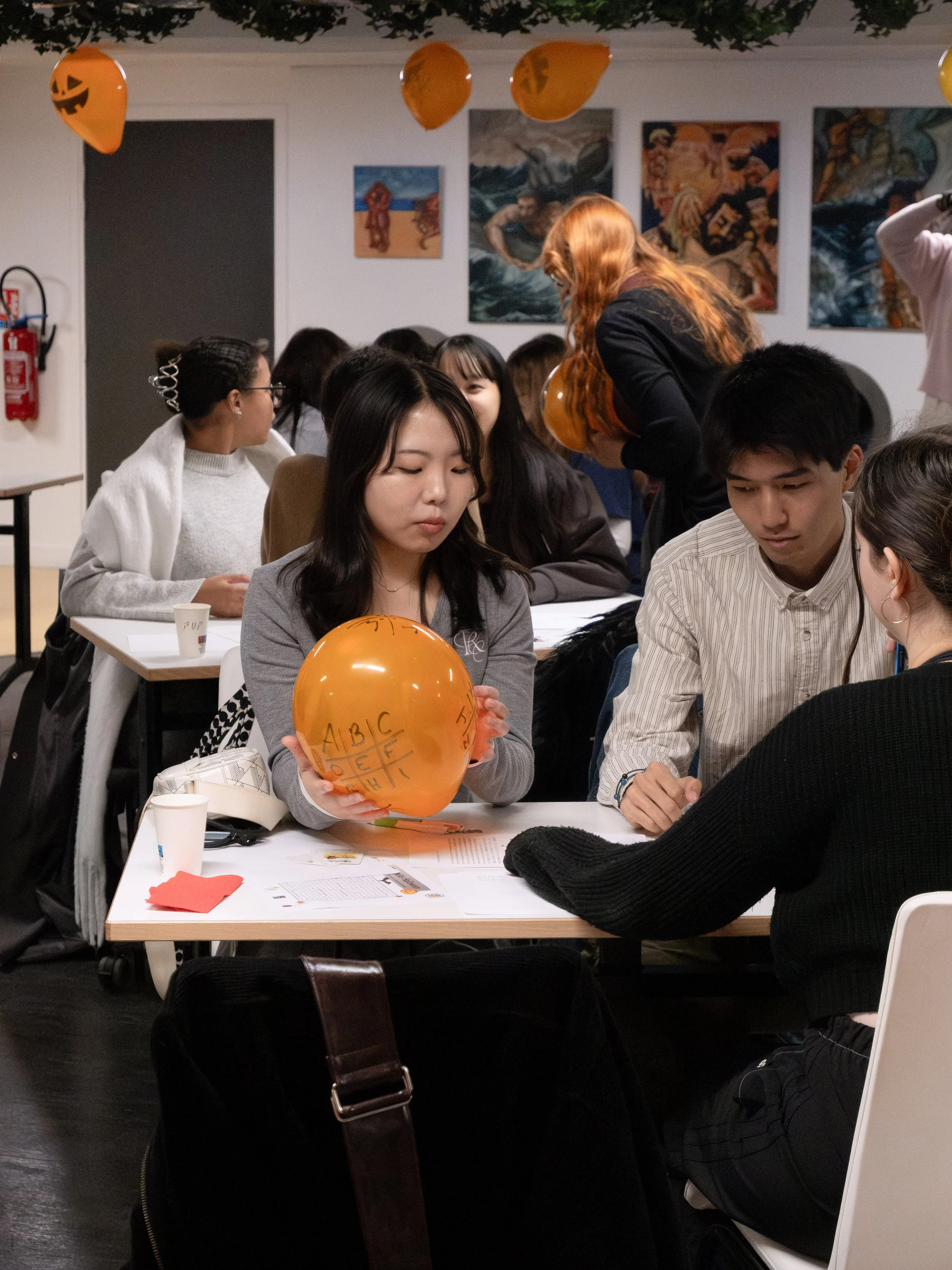 Groupes d'étudiants autour d'une table, certains tenant des ballons orange avec des inscriptions, dans une salle décorée pour Halloween.