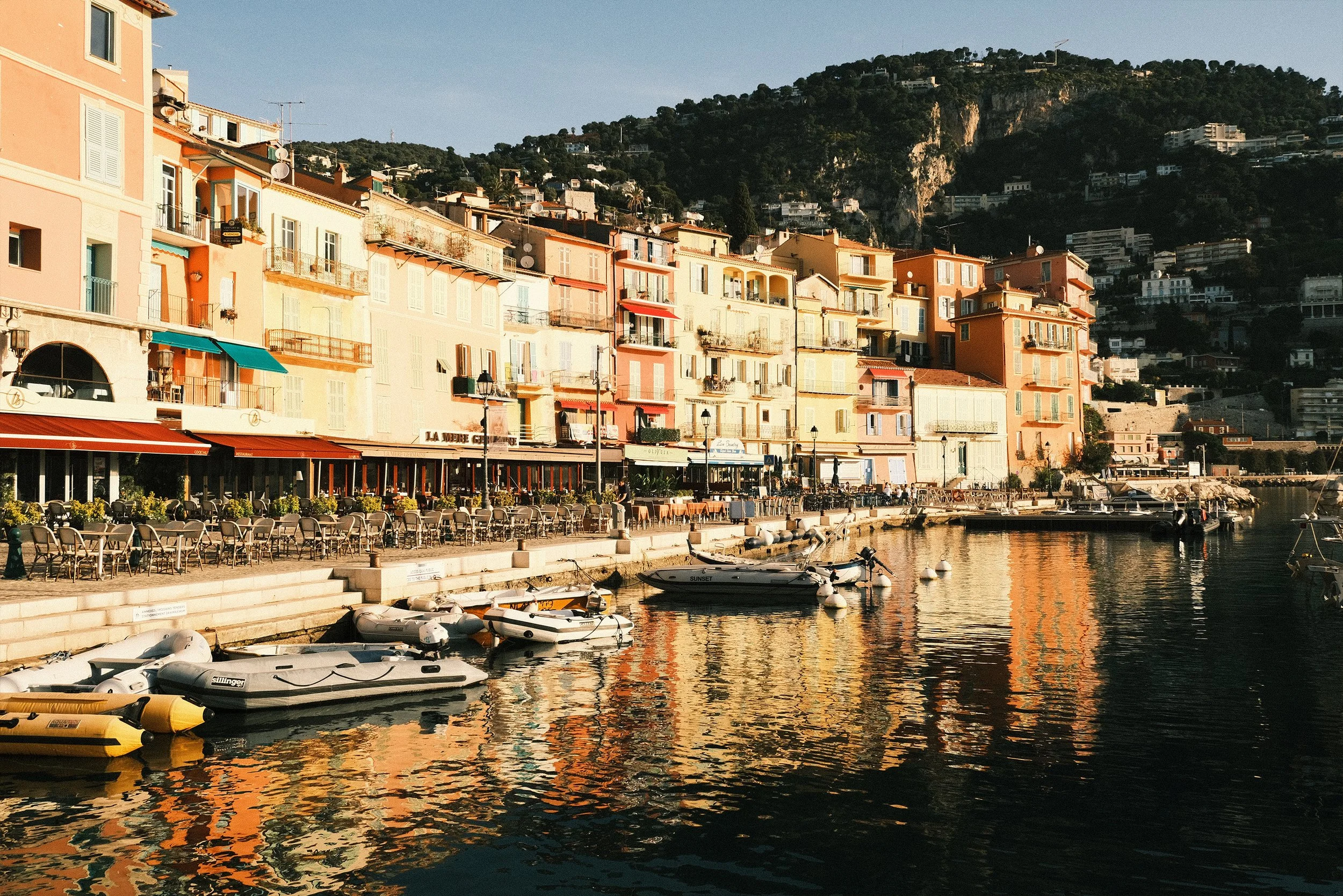 Vue d'un port méditerranéen avec des bateaux amarrés, des maisons colorées de style provençal et des montagnes en arrière-plan, au soleil couchant.