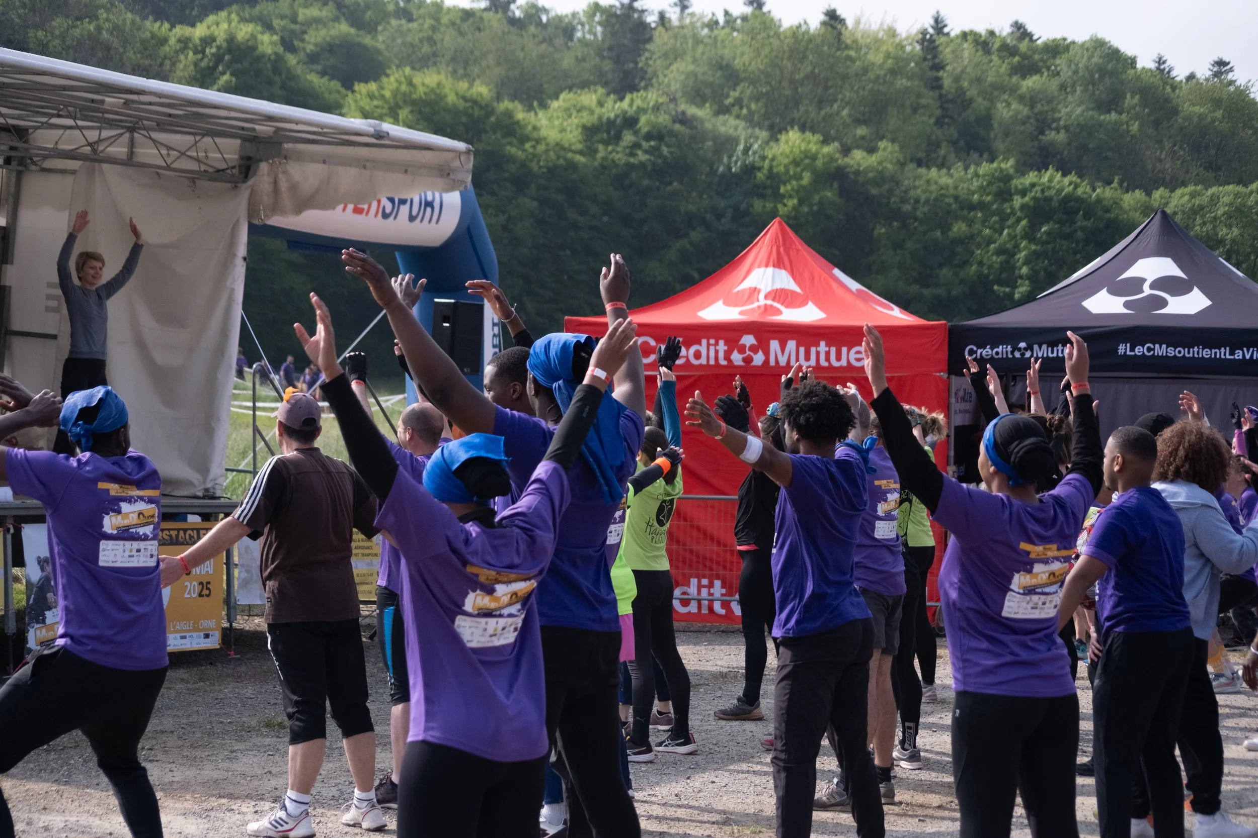 Groupe de personnes, principalement des femmes, participant à une activité de groupe en plein air, portant des T-shirts violets, célébrant ou s'étirant, avec des tentes rouges et noires en arrière-plan portant le nom Crédit Mutuel, dans un environnem