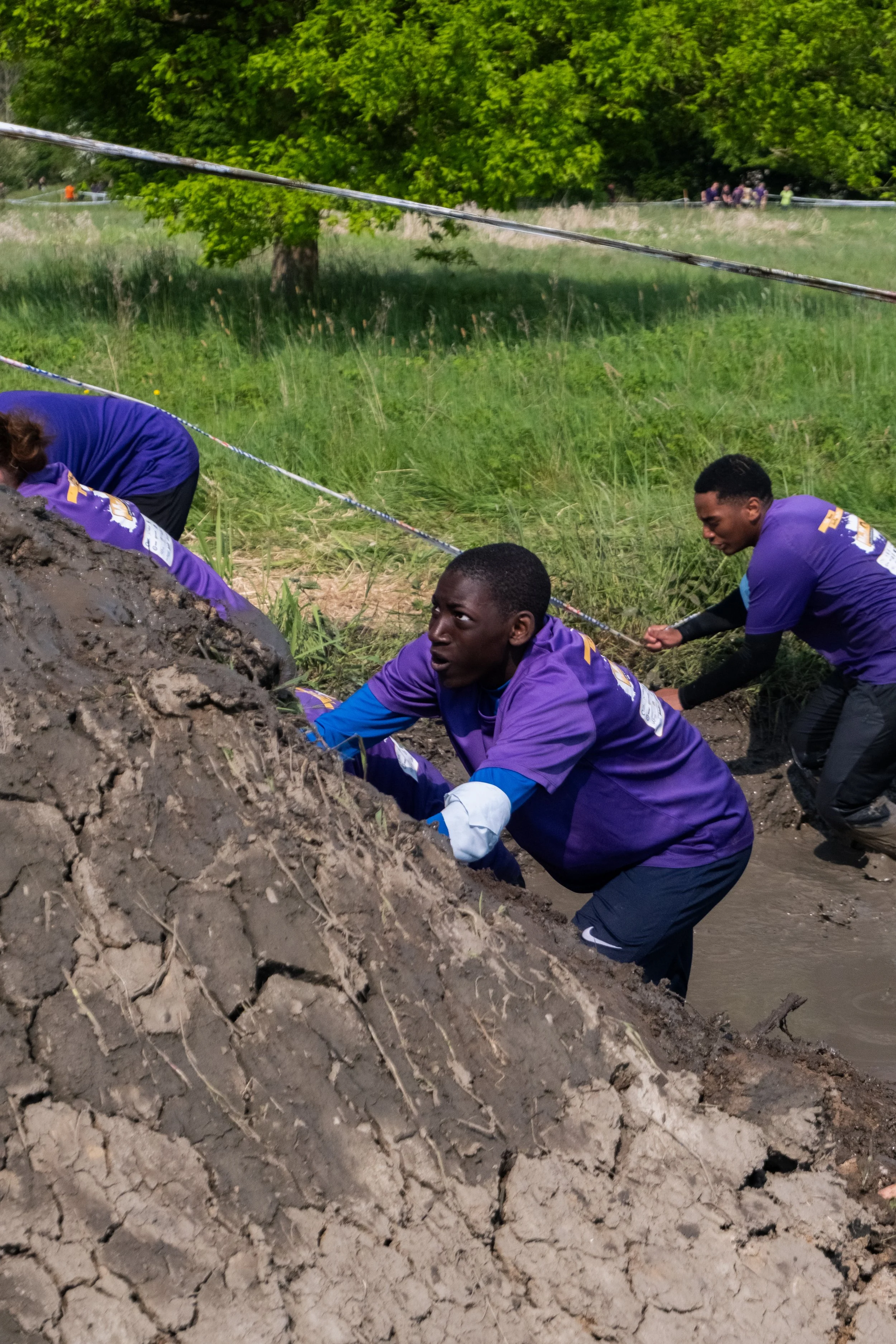 Des jeunes portant des vêtements violets gravissent une pente boueuse lors d'une course d'obstacles en extérieur, avec un terrain verdoyant en arrière-plan.