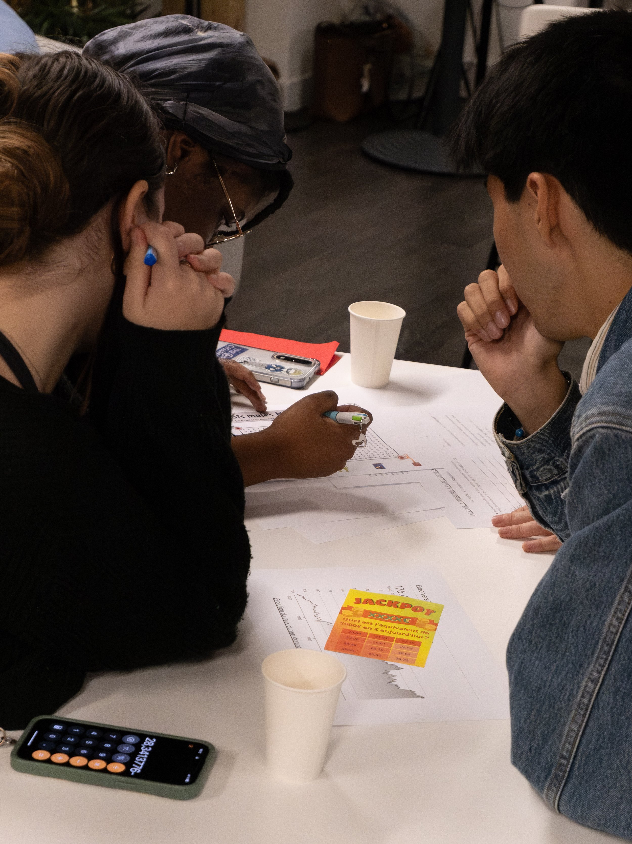 Groupe de trois personnes discutant autour d'une table avec des documents, un téléphone, des gobelets en plastique et une fiche de loterie. La scène se déroule dans une pièce intérieure.