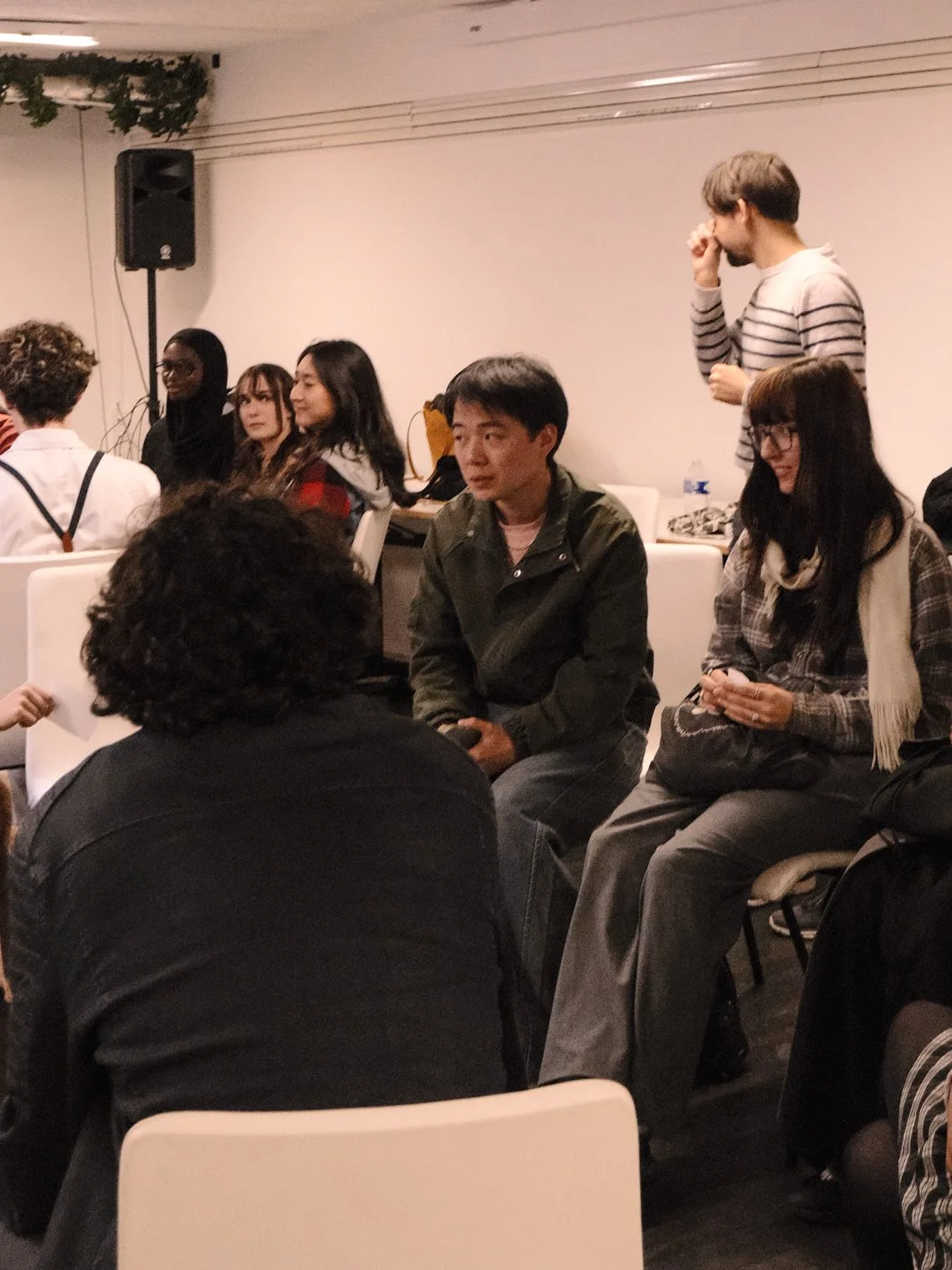 Groupe de personnes assises et discutant dans une salle, avec un homme debout en arrière-plan portant un pull à rayures.