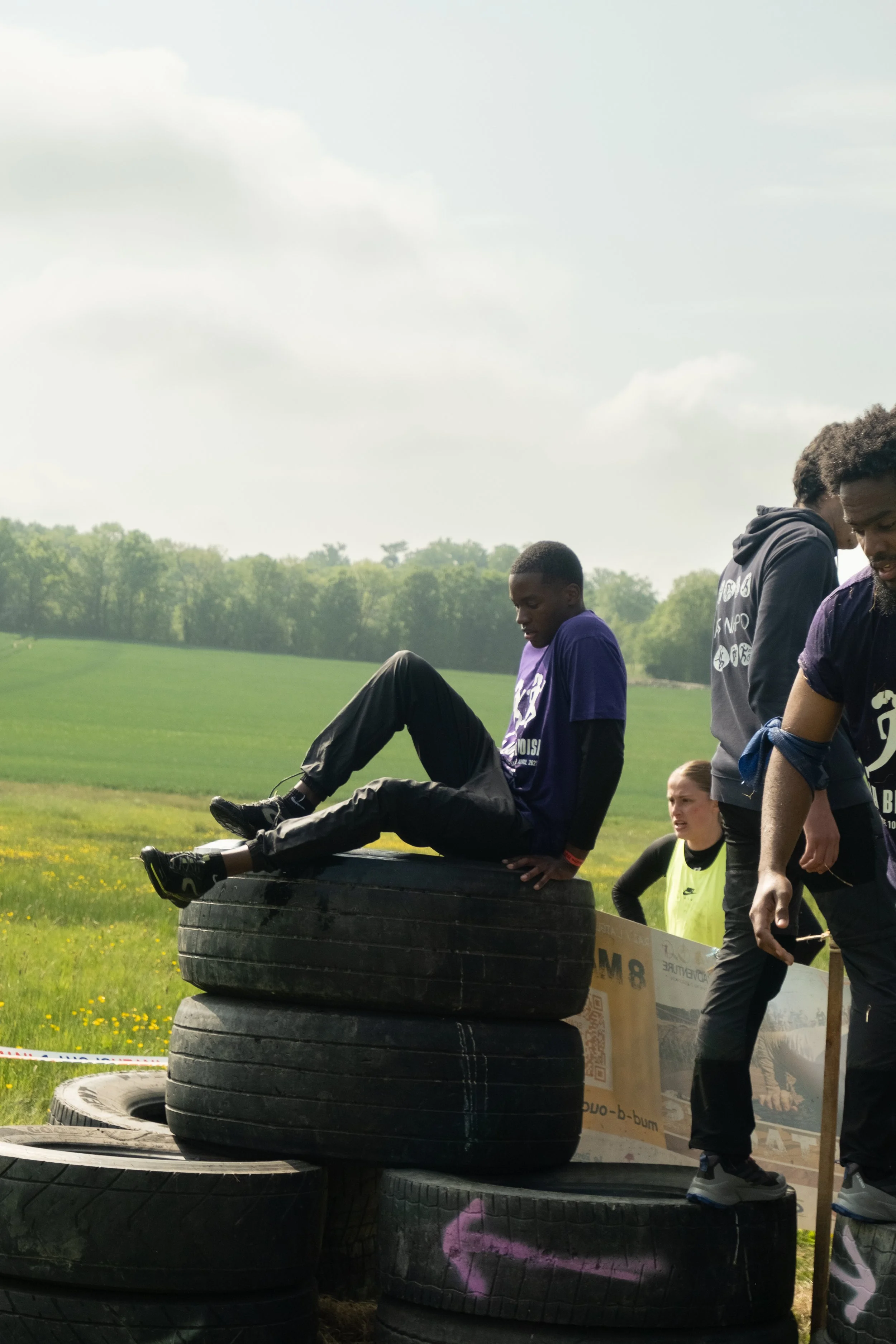 Un groupe de jeunes participe à une activité en extérieur, avec un homme assis sur des pneus empilés dans un champ vert. 