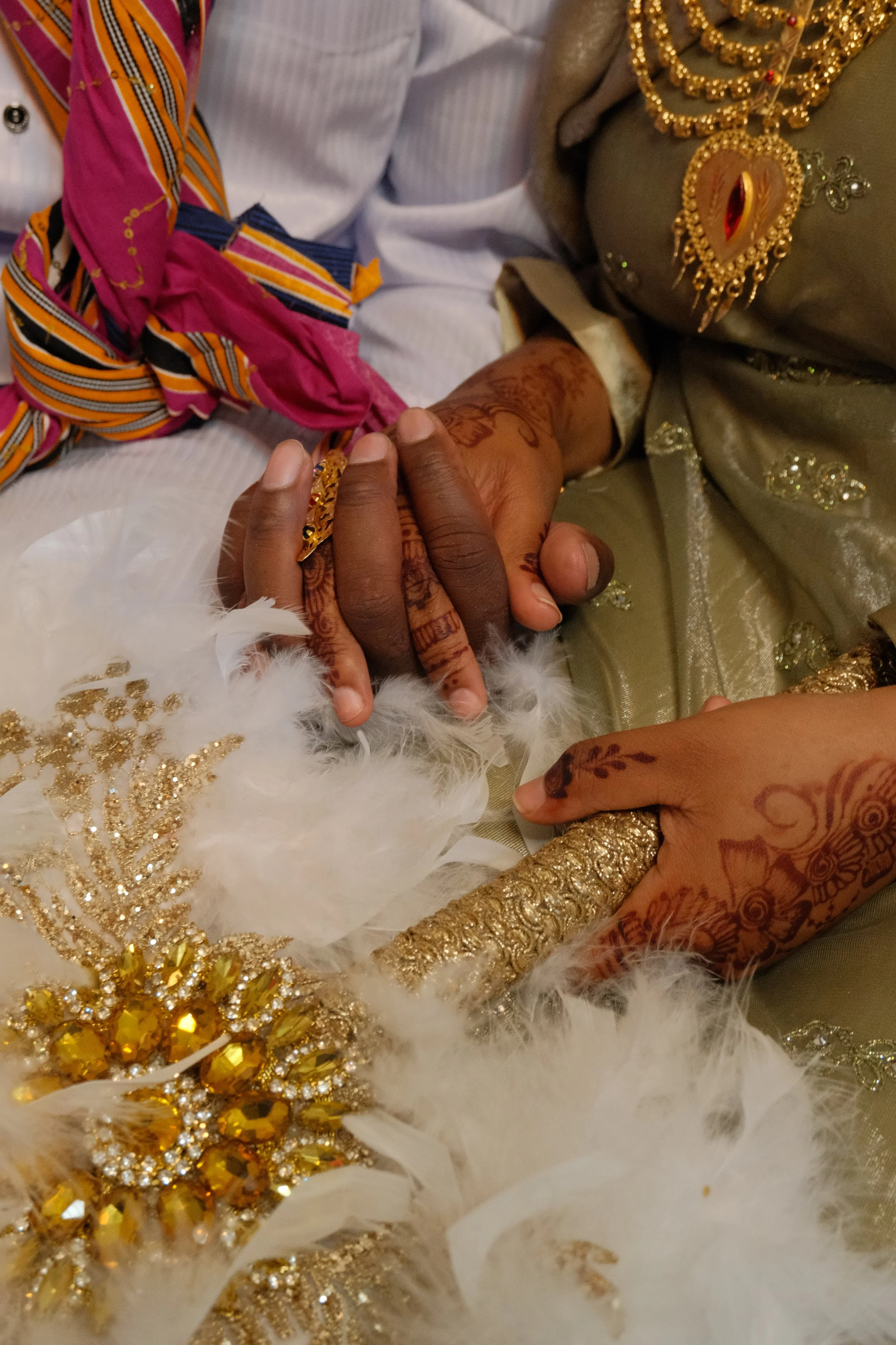 Mains de deux personnes en tenues traditionnelles, décorées de bijoux en or, avec tatouages au henné, tenant un bijou doré et des plumes blanches.
