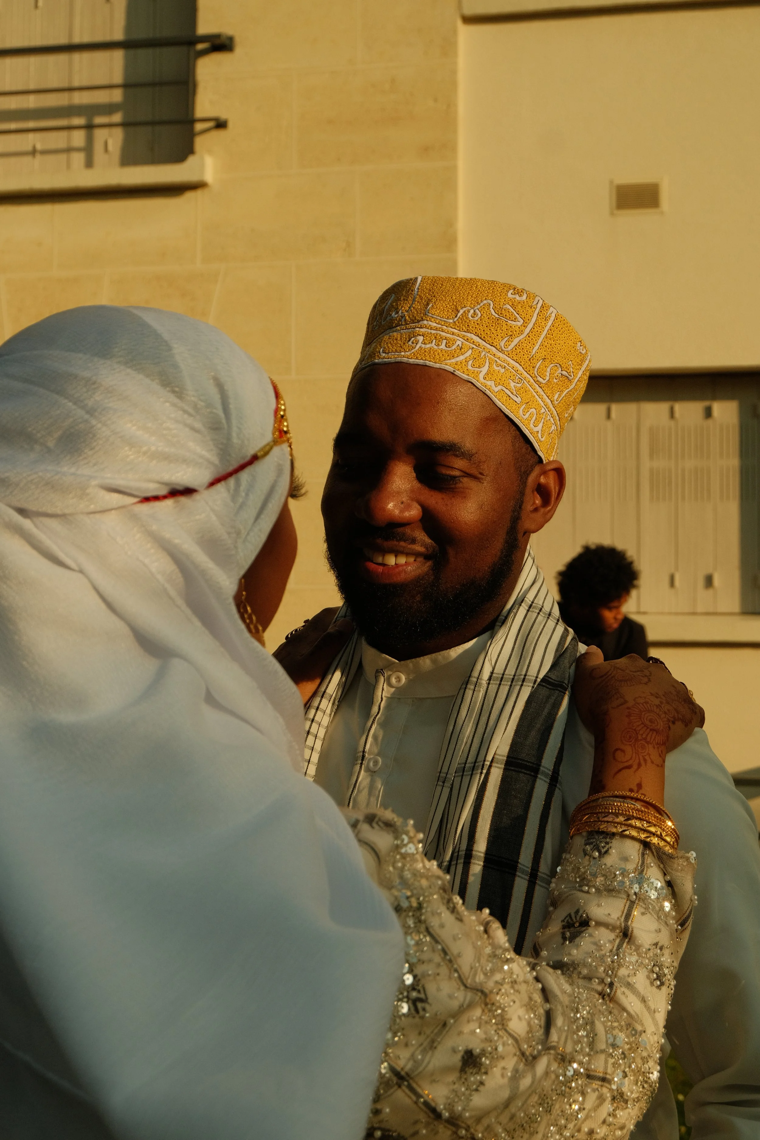 Un homme et une femme en habits traditionnels, partageant un moment d'affection, lors d'une célébration, avec un mur en pierre en arrière-plan.