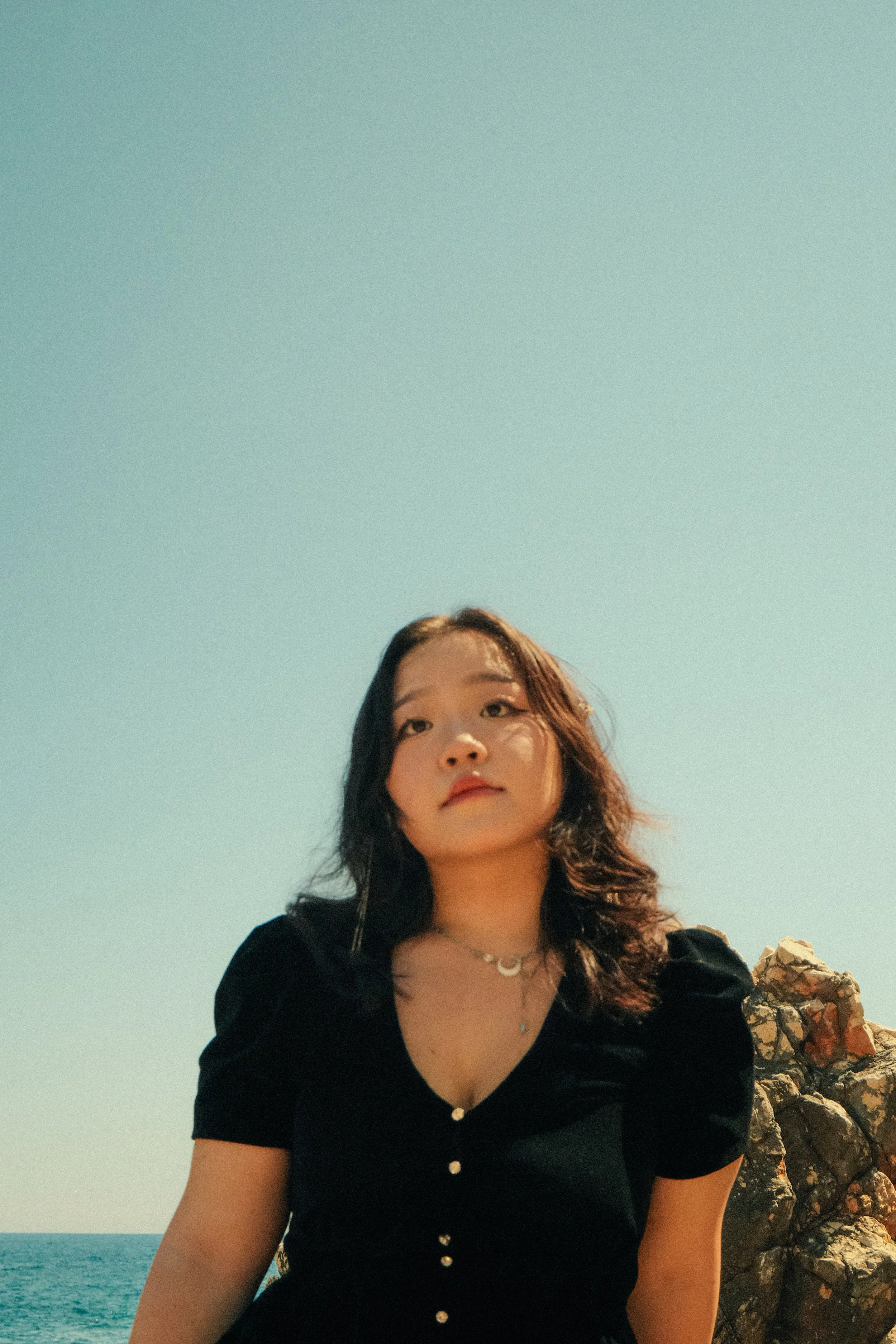 Une jeune femme regardant vers l'objectif, portant une robe noire avec des boutons, avec la mer et un ciel bleu en arrière-plan.