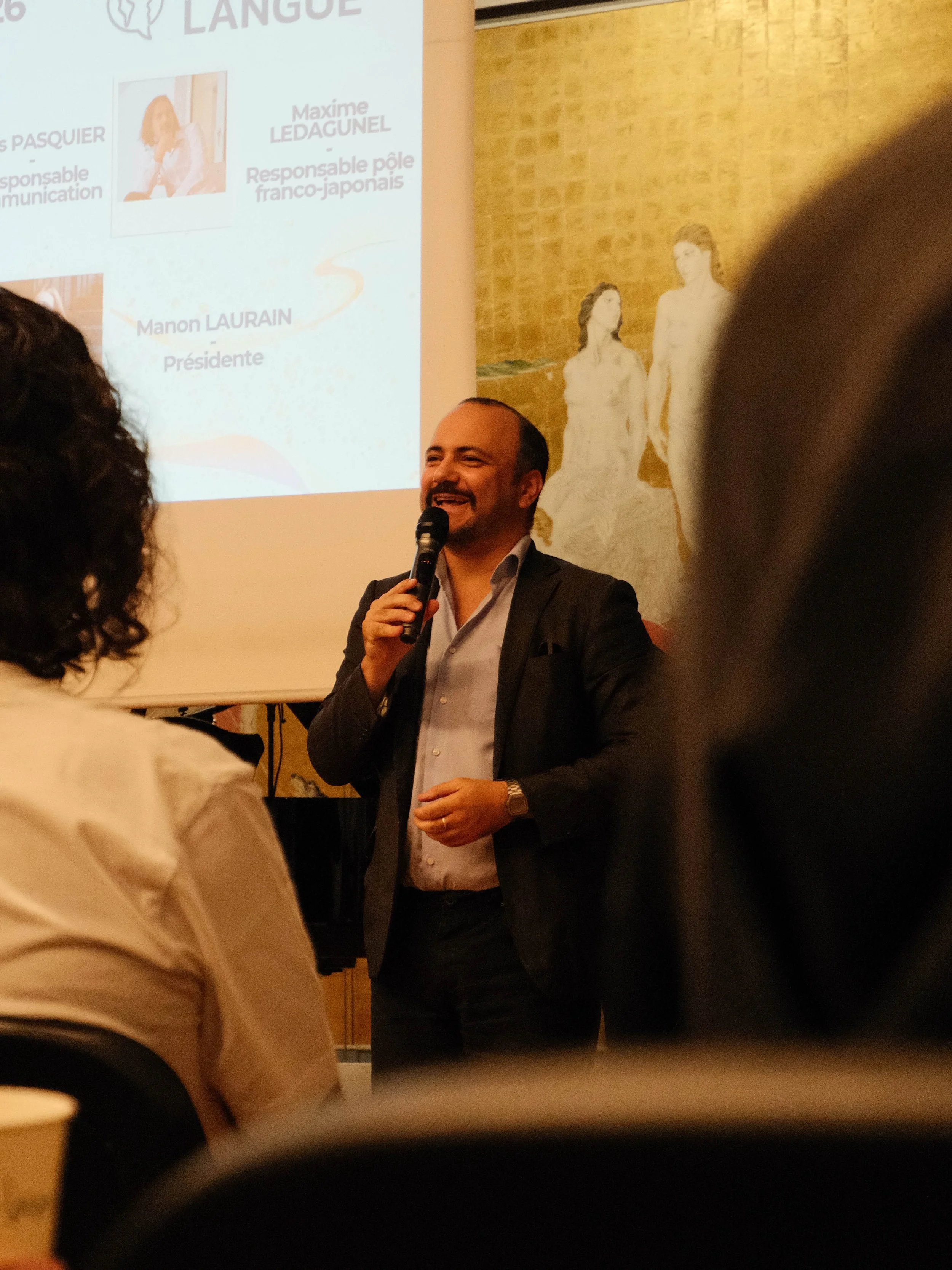 Un homme souriant tient un micro lors d'une présentation, avec un écran projeté en arrière présentant des noms et rôles. La scène est intérieure avec un fond en briques.