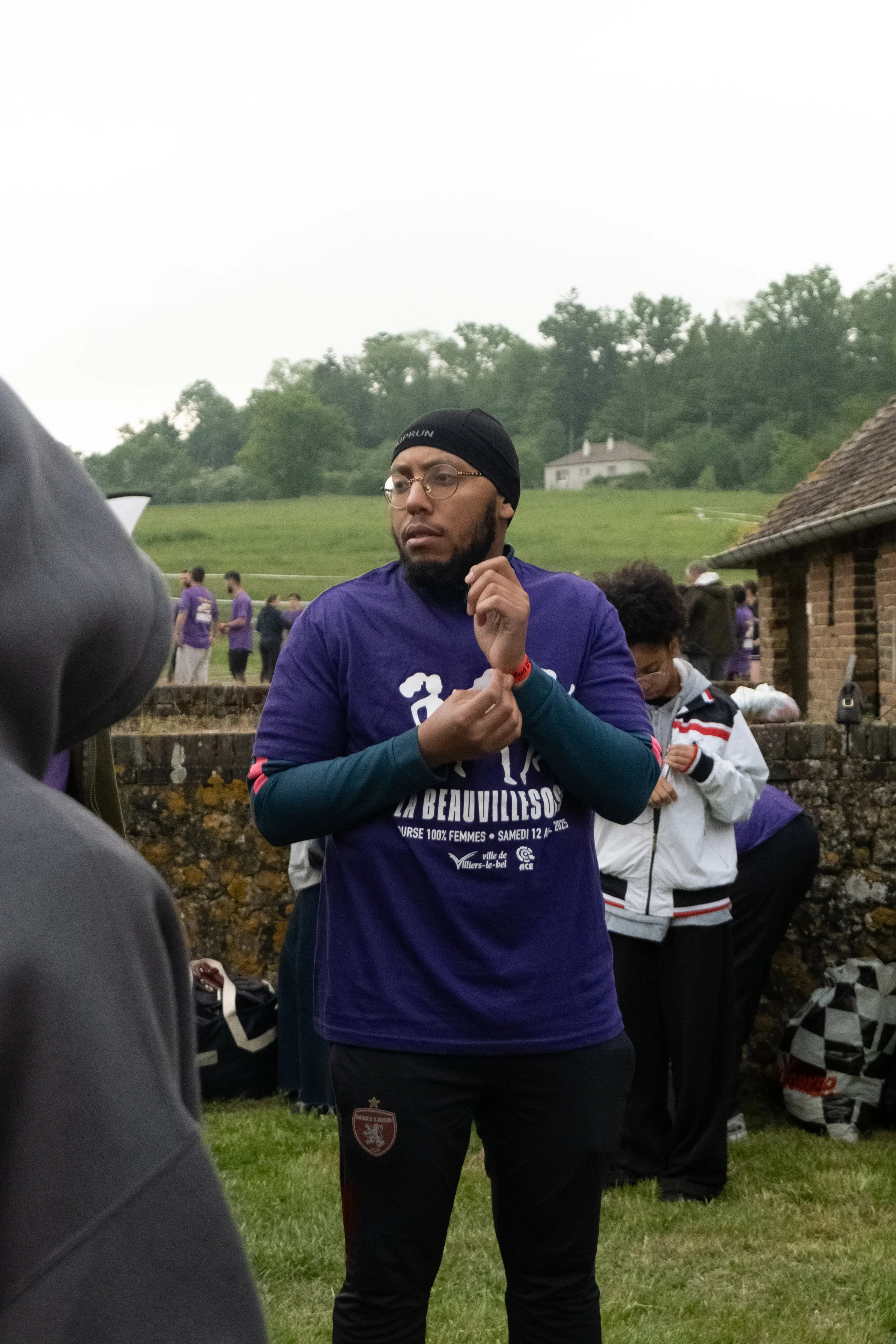 Un homme portant un bandeau noir, des lunettes, un t-shirt violet avec des inscriptions en français participe à un événement en plein air avec d'autres personnes en arrière-plan, dans un cadre rural avec une maison en pierre et un paysage verdoyant.