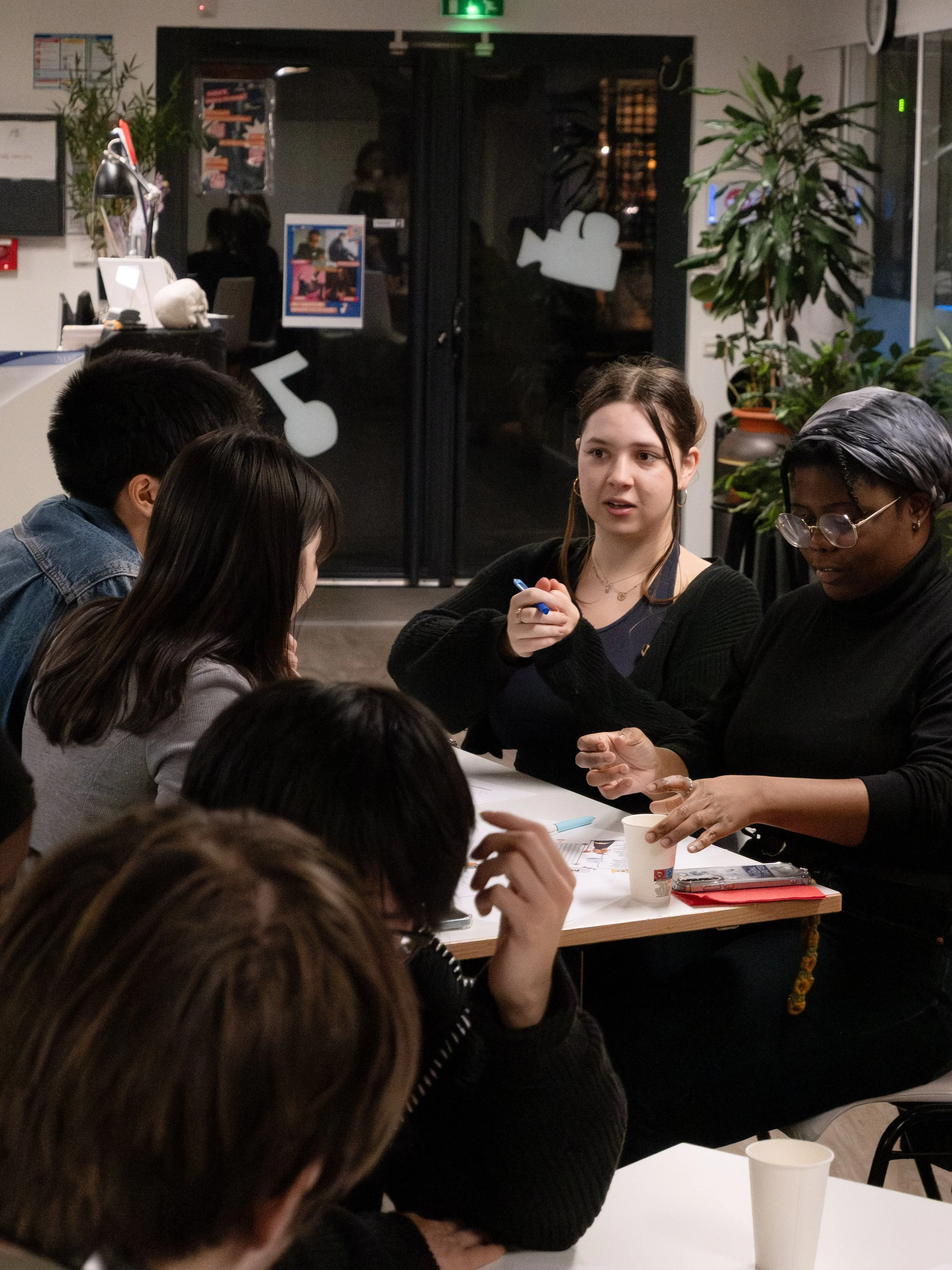 Groupe de personnes discutant autour d'une table dans un café ou un espace de réunion, avec des plantes en arrière-plan et une porte vitrée dans la pièce.
