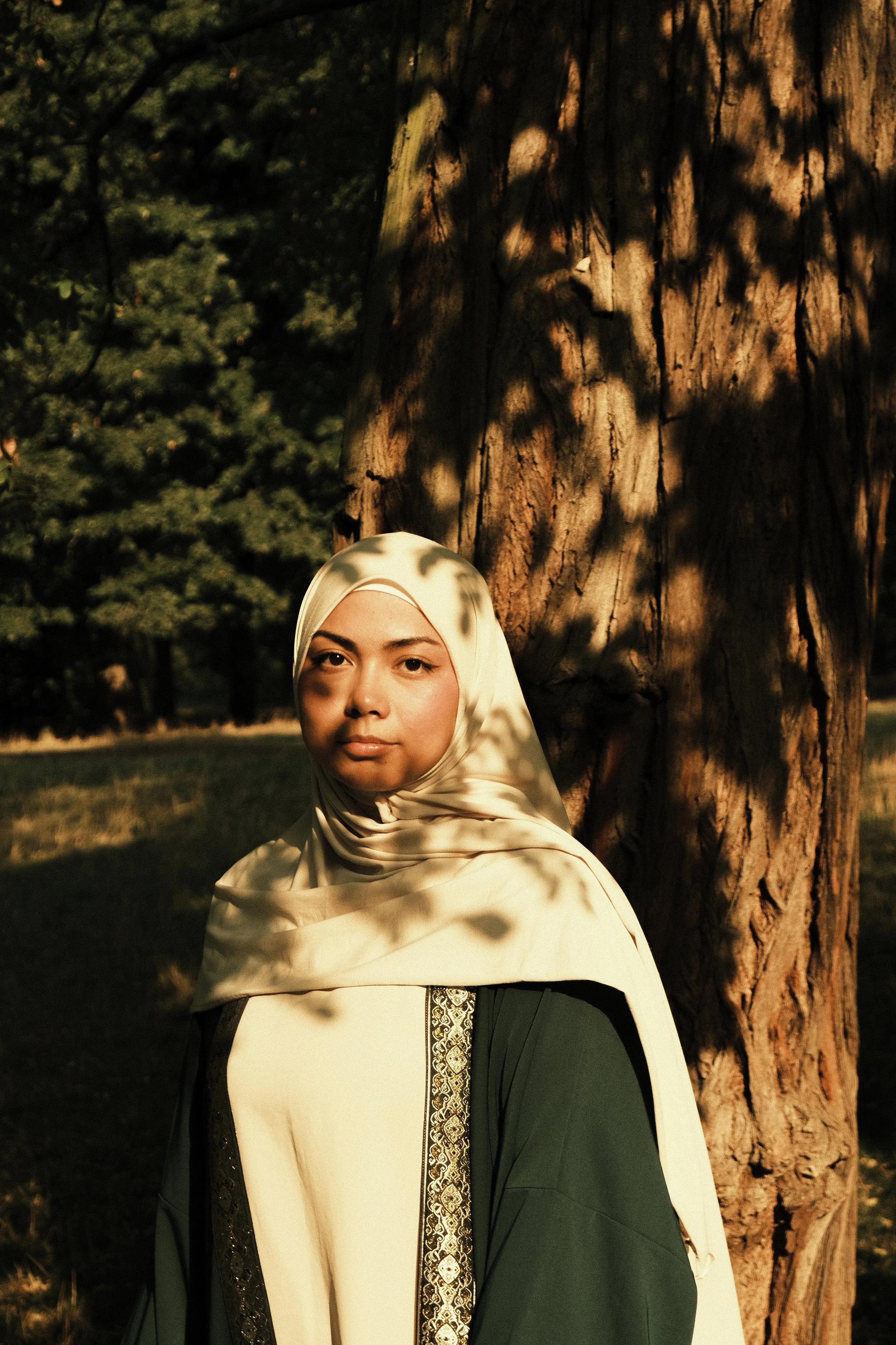 Une femme portant un hijab crème et une robe verte, se tenant devant un arbre en plein air, avec des ombres de feuilles projetées sur son visage et ses épaules, dans une lumière dorée