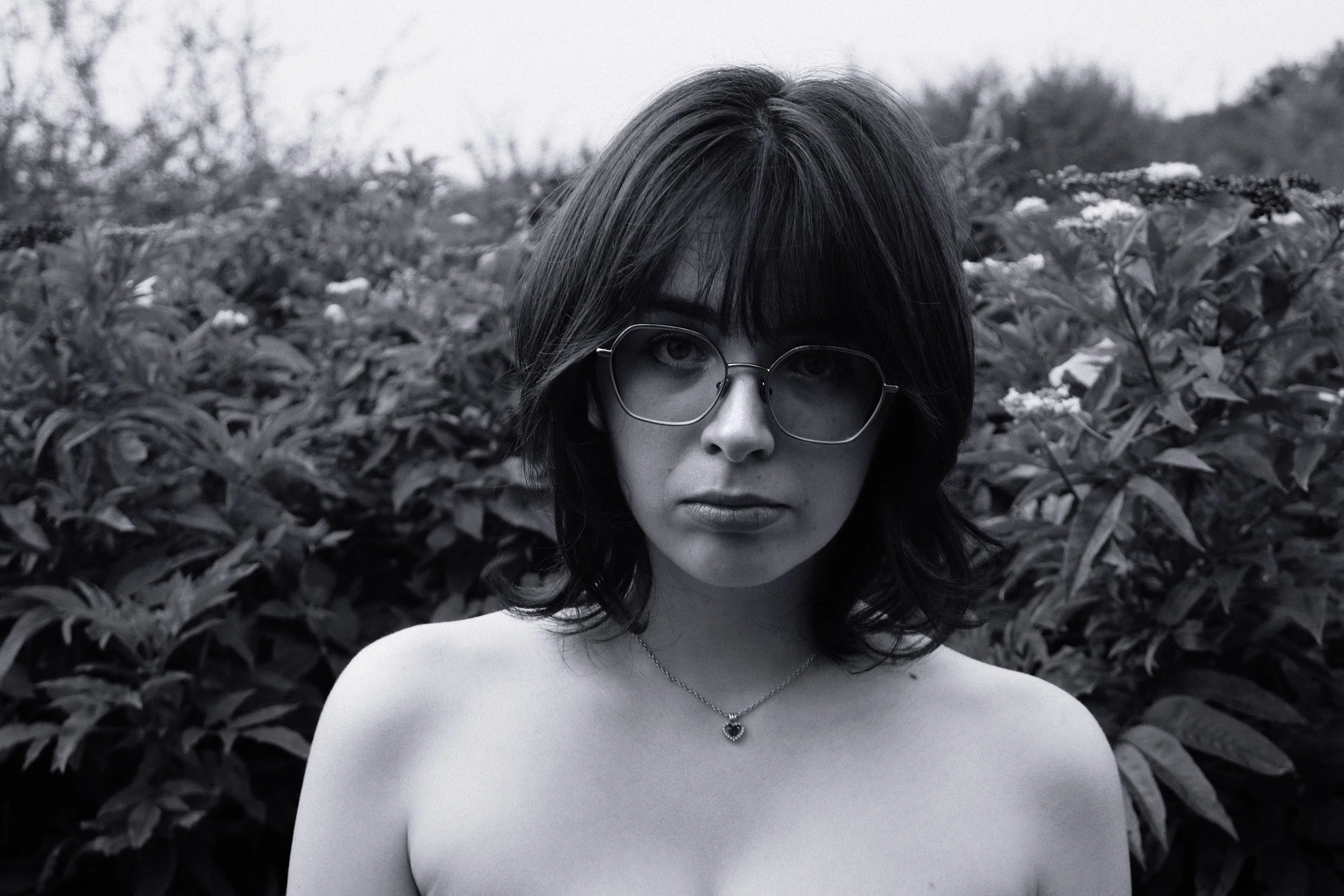 Portrait en noir et blanc d'une femme aux cheveux courts, portant des lunettes rondes et un collier avec un pendentif cœur, dans un champ de végétation.