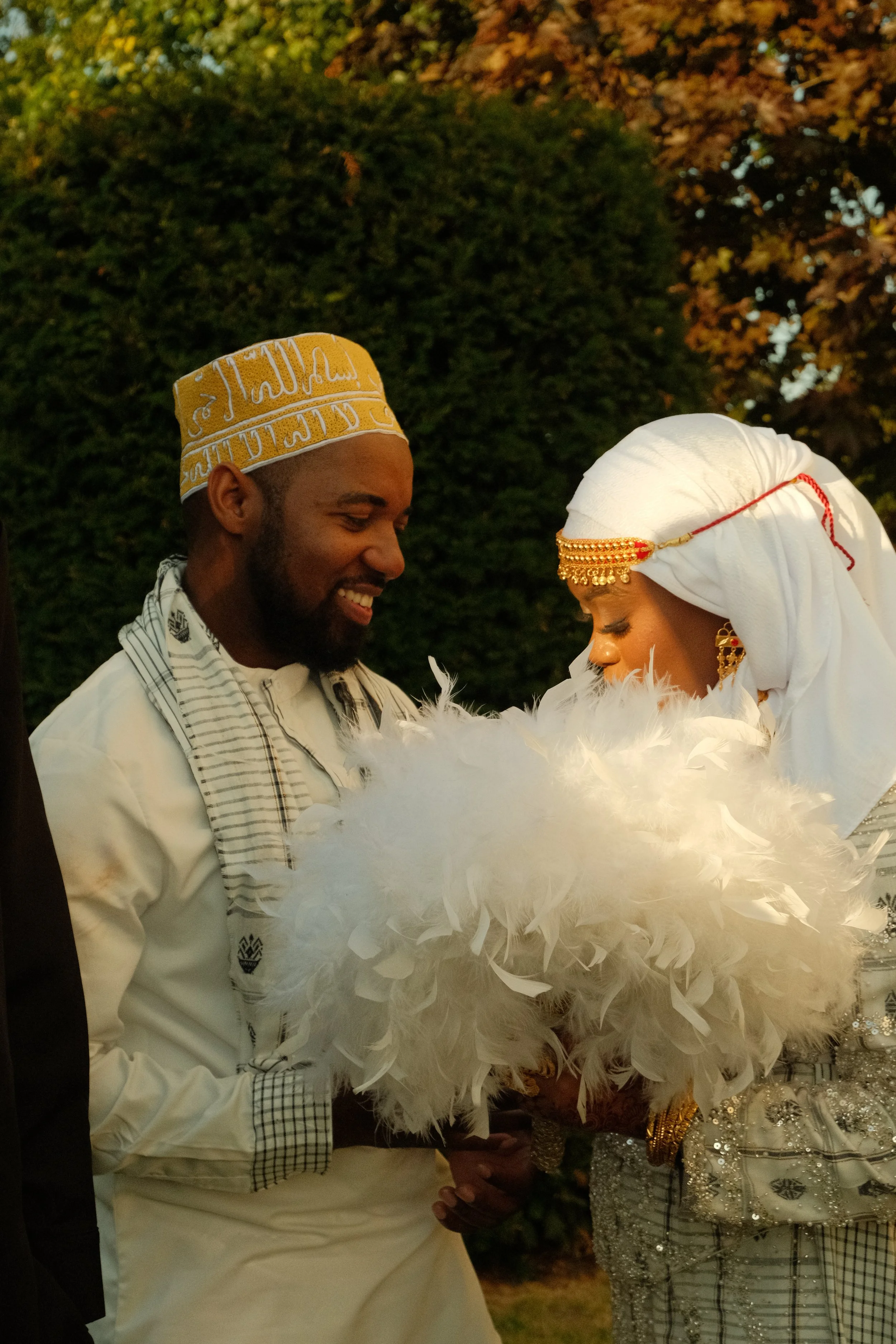Un homme et une femme lors d'une cérémonie, la femme porte un tissu blanc avec des accessoires dorés et un voile, elle tient un bouquet blanc en plumes, ils se tiennent la main et semblent échanger un regard, dans un décor naturel avec des arbres.