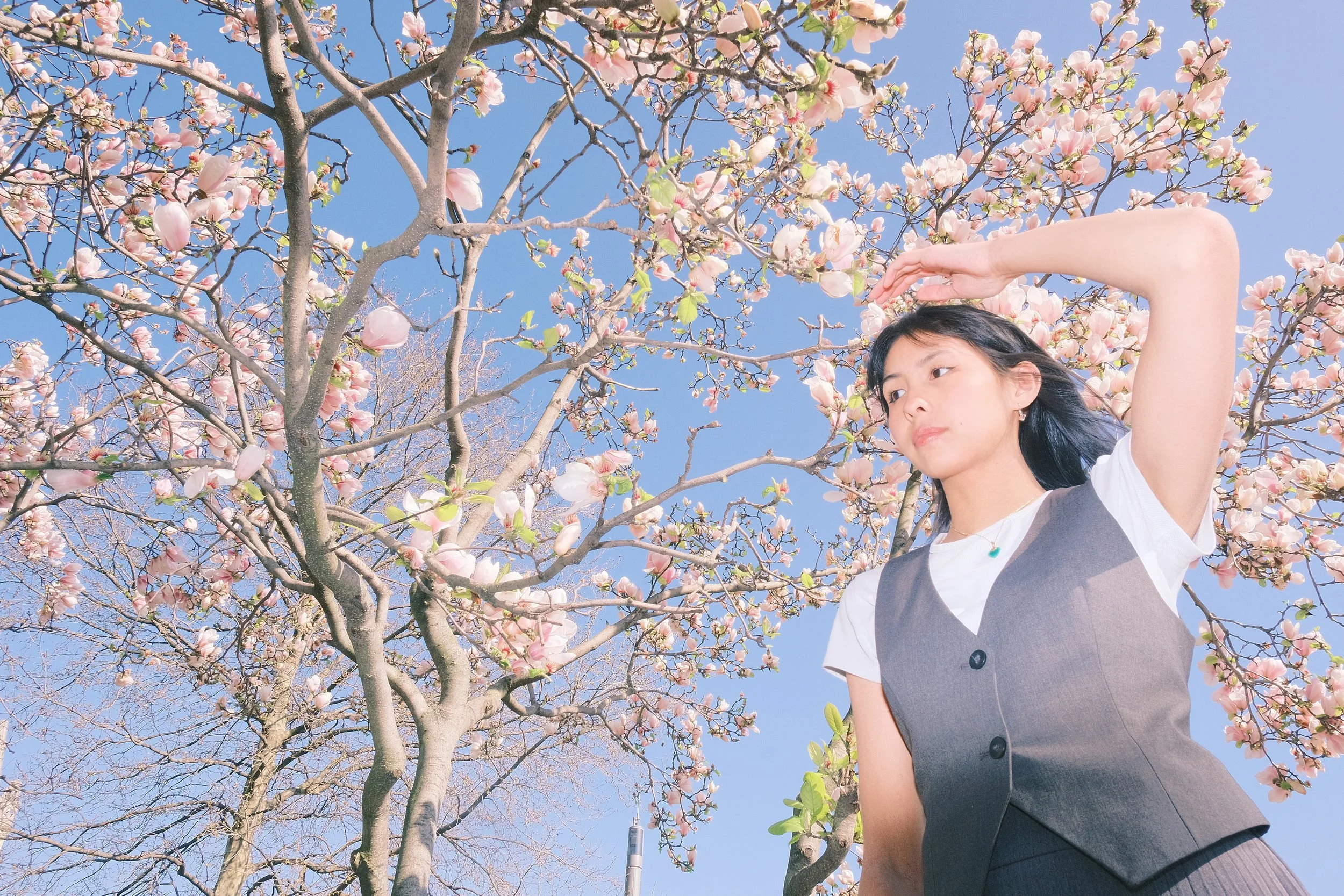 Jeune femme avec une robe grise et un t-shirt blanc, se tenant devant un arbre en fleurs roses contre un ciel bleu, en train de se protéger du soleil avec la main droite