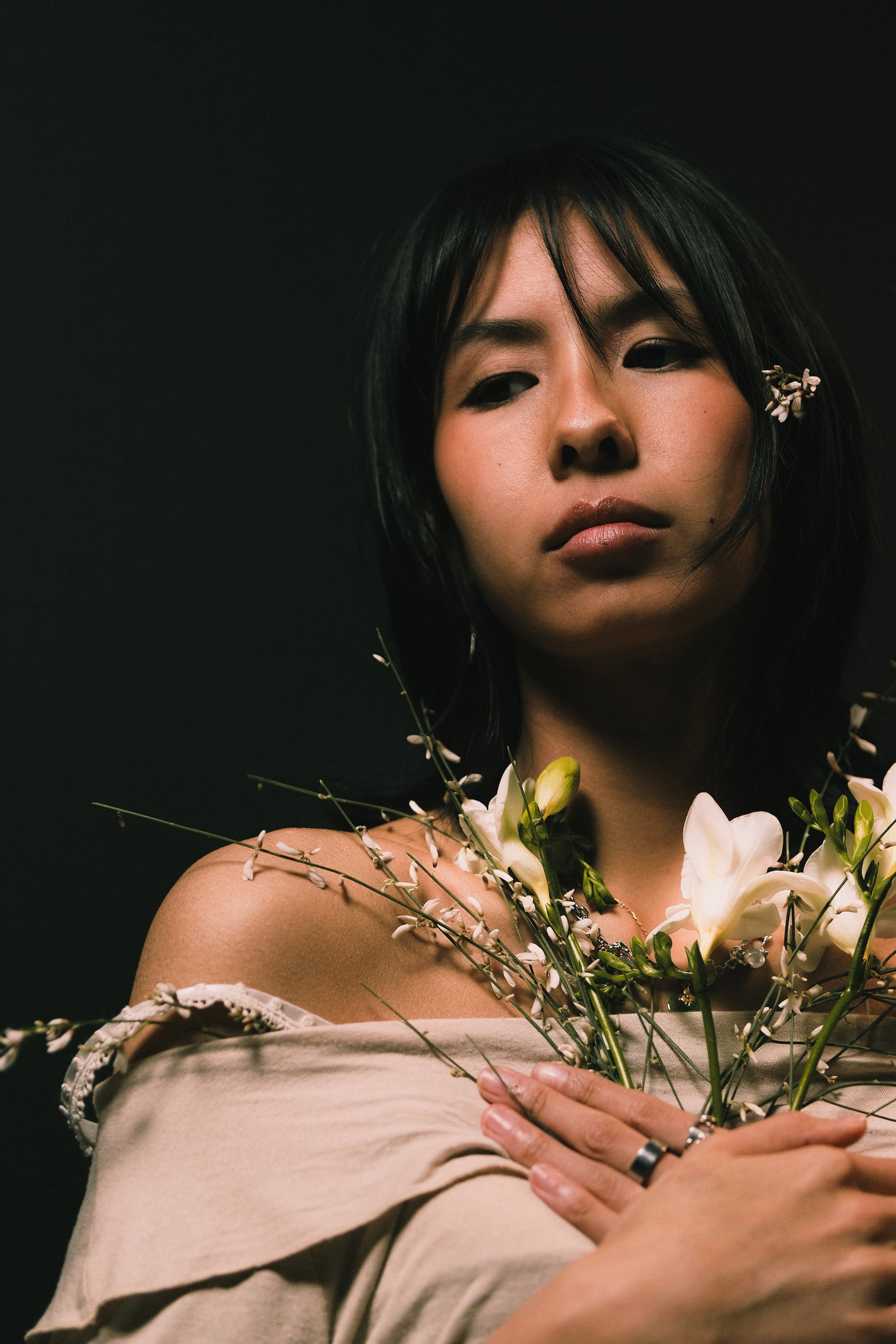 Jeune femme asiatique avec des fleurs nues. Elle tient un bouquet de fleurs blanches contre sa poitrine et regarde fixement devant elle, avec une expression sérieuse.