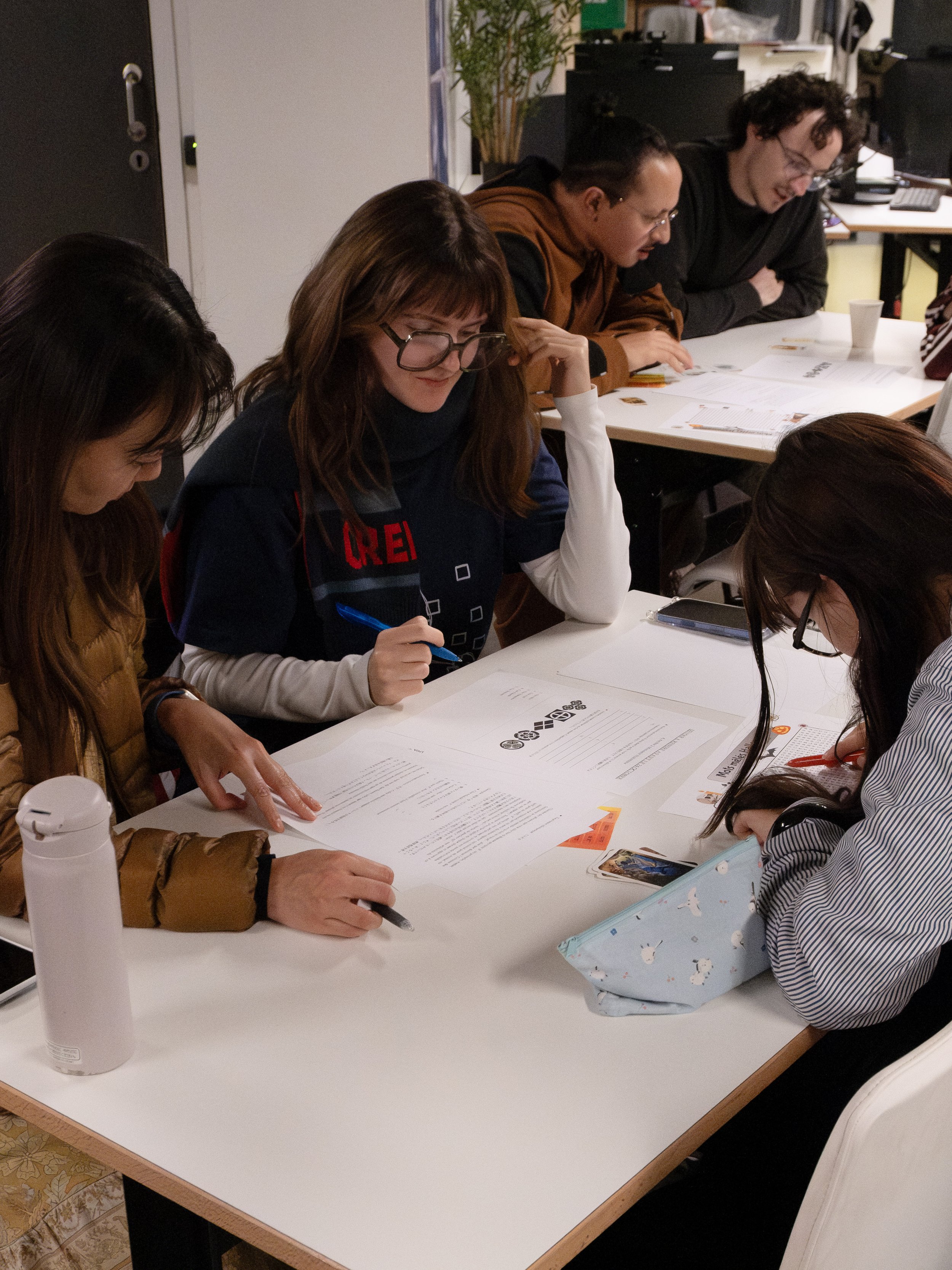 Groupe de personnes assises autour d'une table en train de lire et d'écrire, dans un environnement intérieur, probablement une salle de réunion ou une classe.