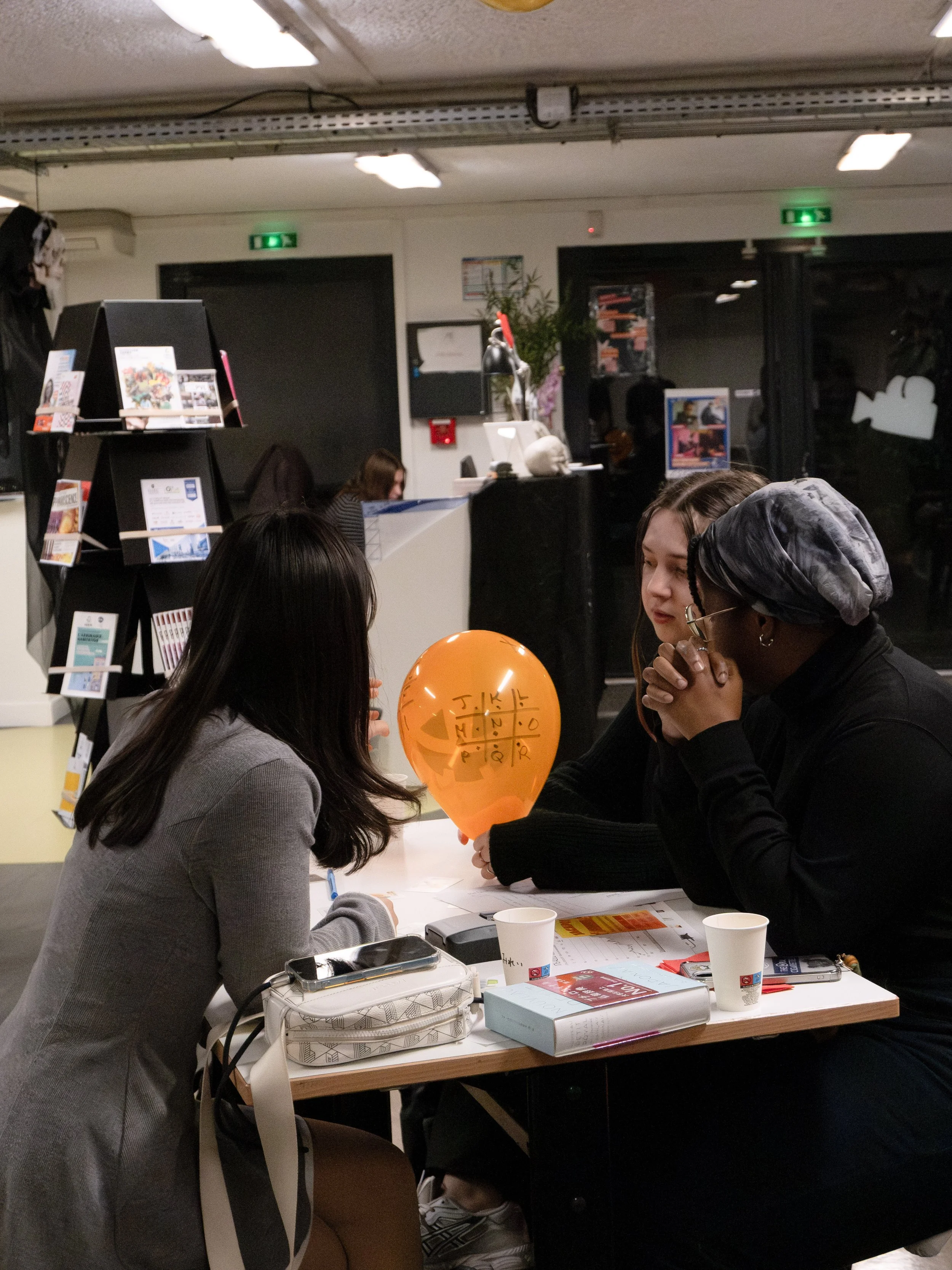 Trois femmes assises autour d'une table discutant, avec un ballon orange portant une grille de mots croisés anglais, dans un café ou un lieu public.