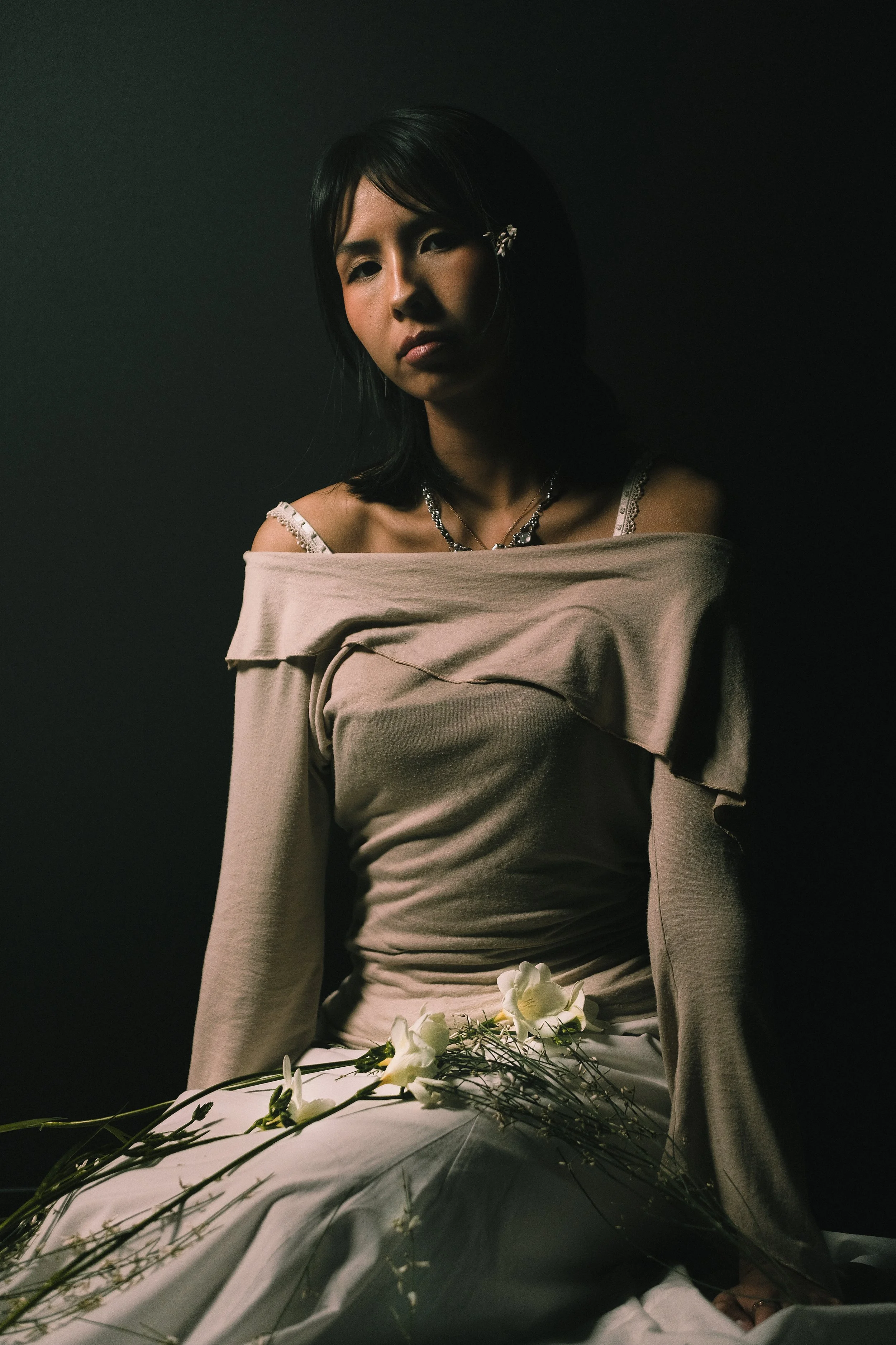 Une femme avec des vêtements beige, portant des bijoux, assise dans un environnement sombre, avec des fleurs blanches devant elle.