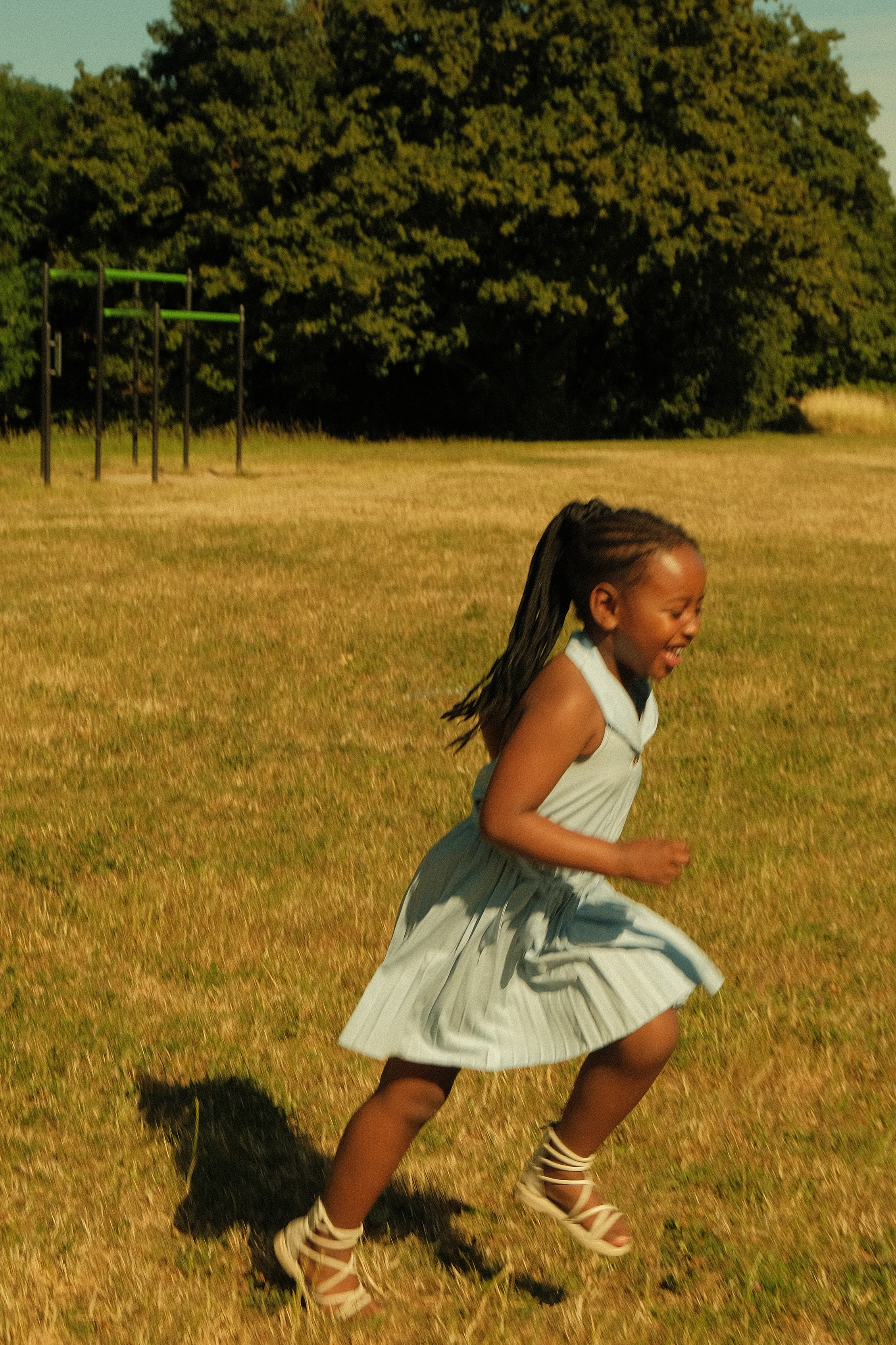 Une jeune fille souriante qui court dans un jardin ensoleillé avec un arbre et une structure de jeu en arrière-plan.