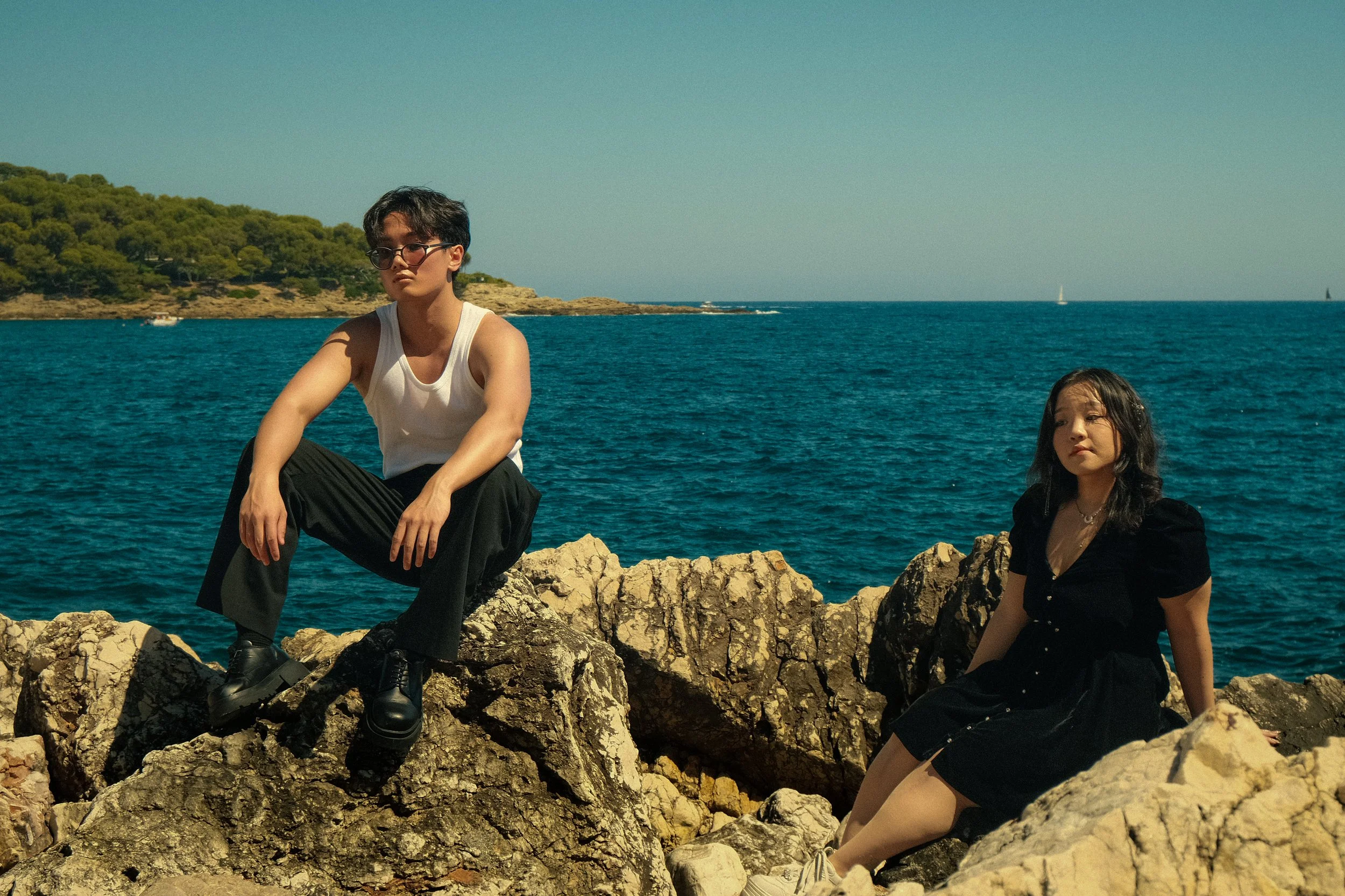 Deux jeunes femmes assises sur des rochers au bord de la mer, avec un paysage marin et un ciel bleu en arrière-plan.