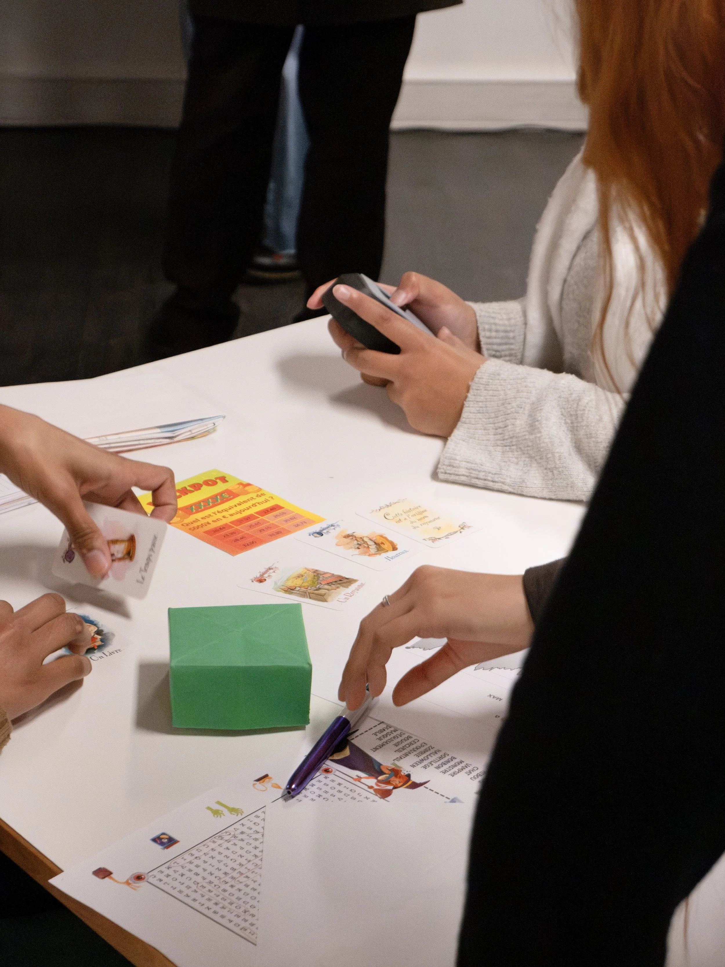 Groupe de personnes jouant à un jeu de société autour d'une table, avec divers papiers, un stylo, des cartes et un cube en papier vert.